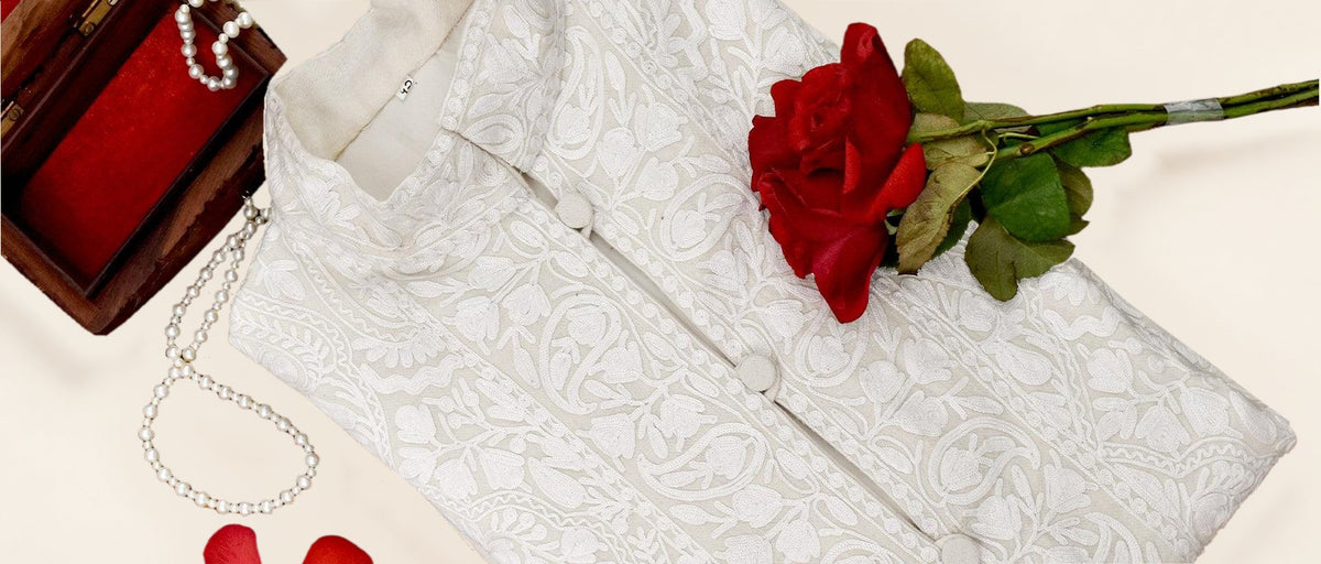 Folded ivory-white Aari embroidered jacket arranged with red roses and pearl strands on a patterned surface