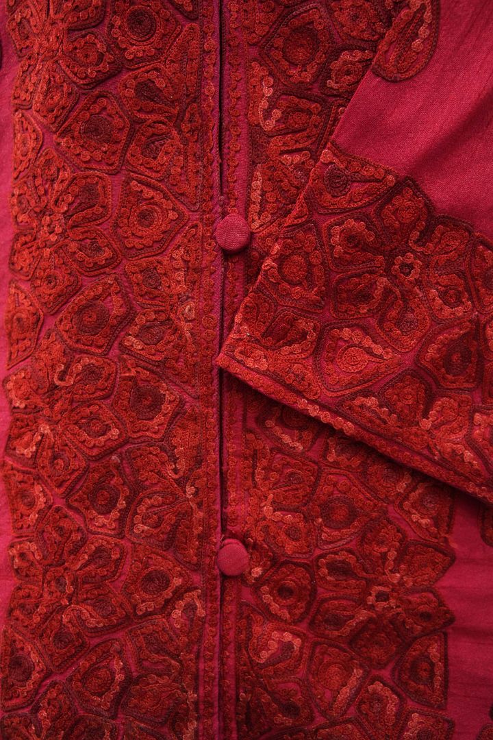 Detail of a folded ruby-red cuff against the buttoned front, both densely decorated with tonal Aari floral and paisley motifs.