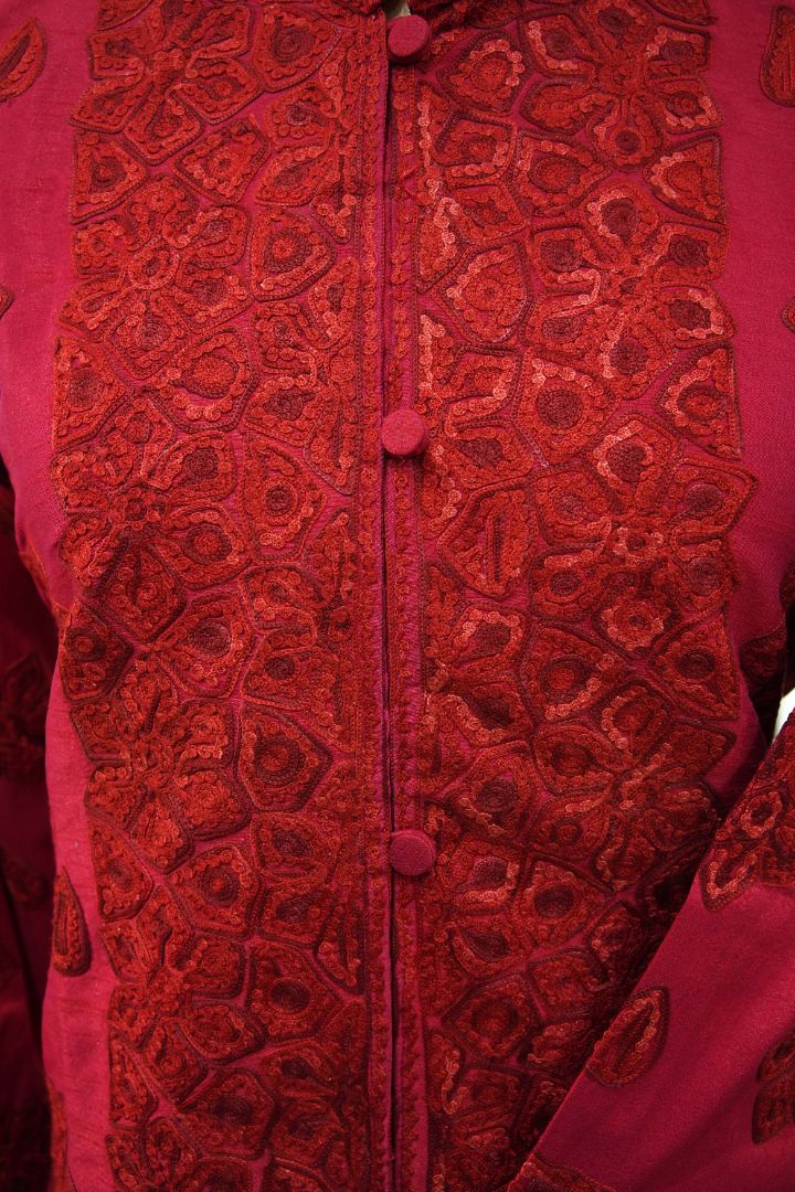Close-up of the ruby-red jacket’s buttoned front, with small Aari floral and paisley motifs arranged in irregular outlined panels.