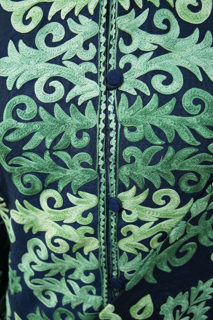 Close-up of green Aari leaf scrolls flanking the navy jacket’s covered buttons and narrow embroidered opening