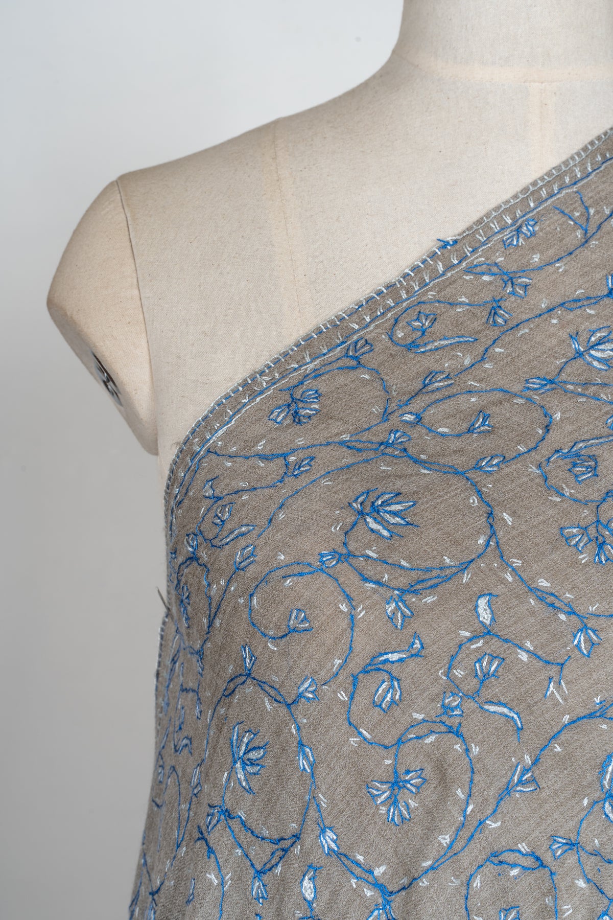 Close-up of sky-blue and pale Sozni flowers on curling vines across a grey shawl at the mannequin's shoulder