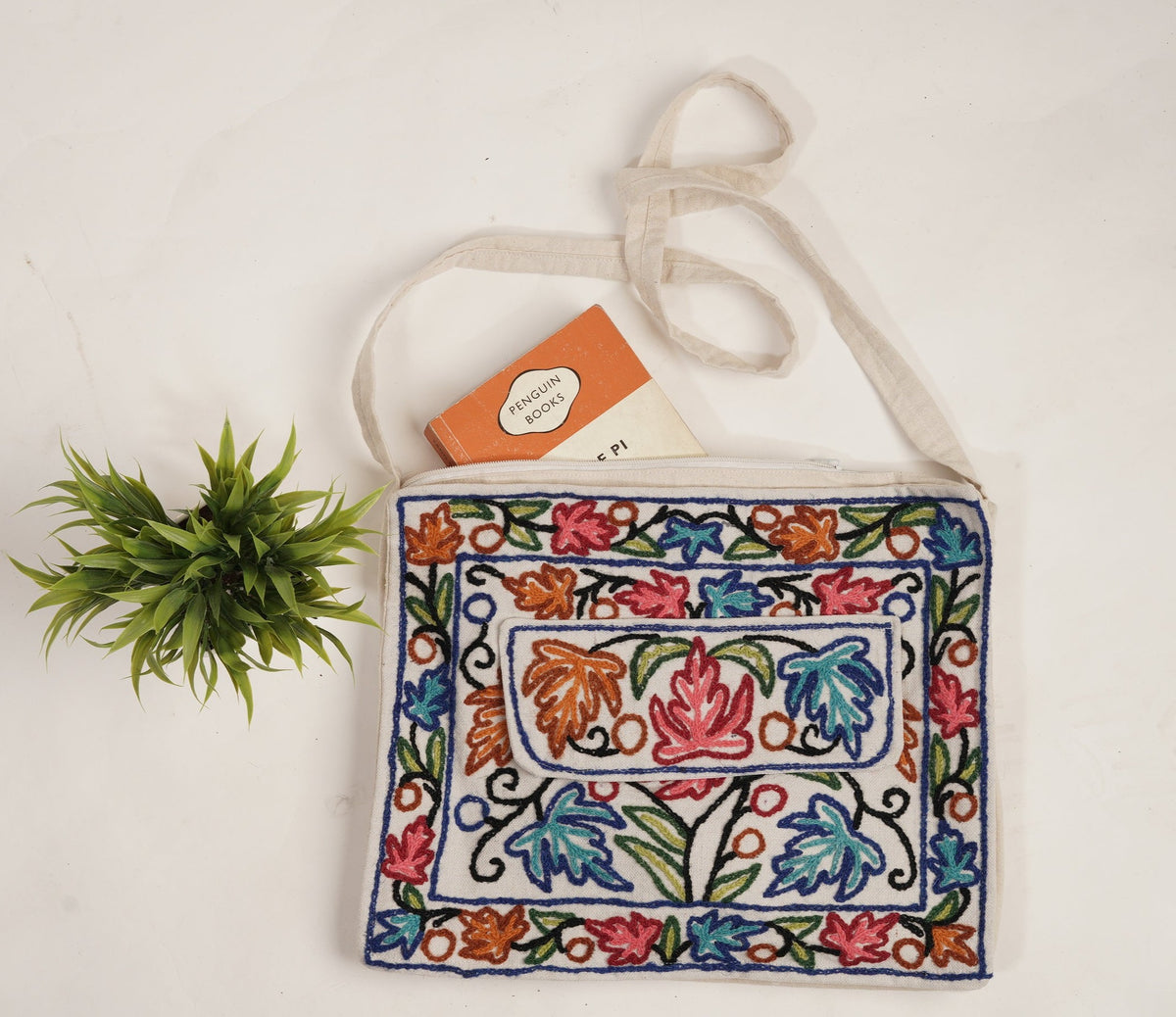 Cream cotton shoulder bag laid flat, with red, blue, green and orange Aari floral embroidery, navy outlines and a long cream strap