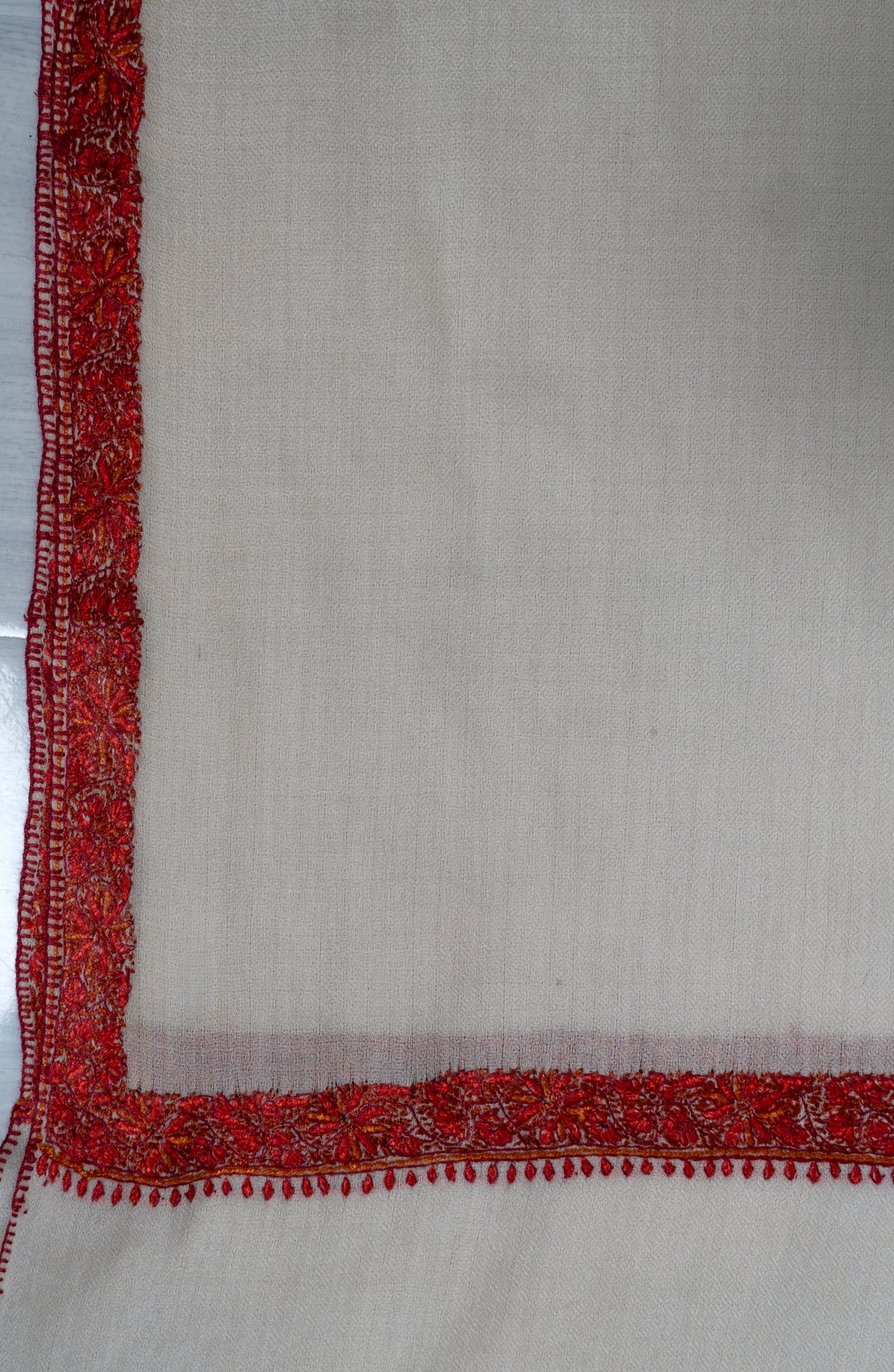 Flat corner detail of an ivory shawl framed by narrow crimson floral embroidery