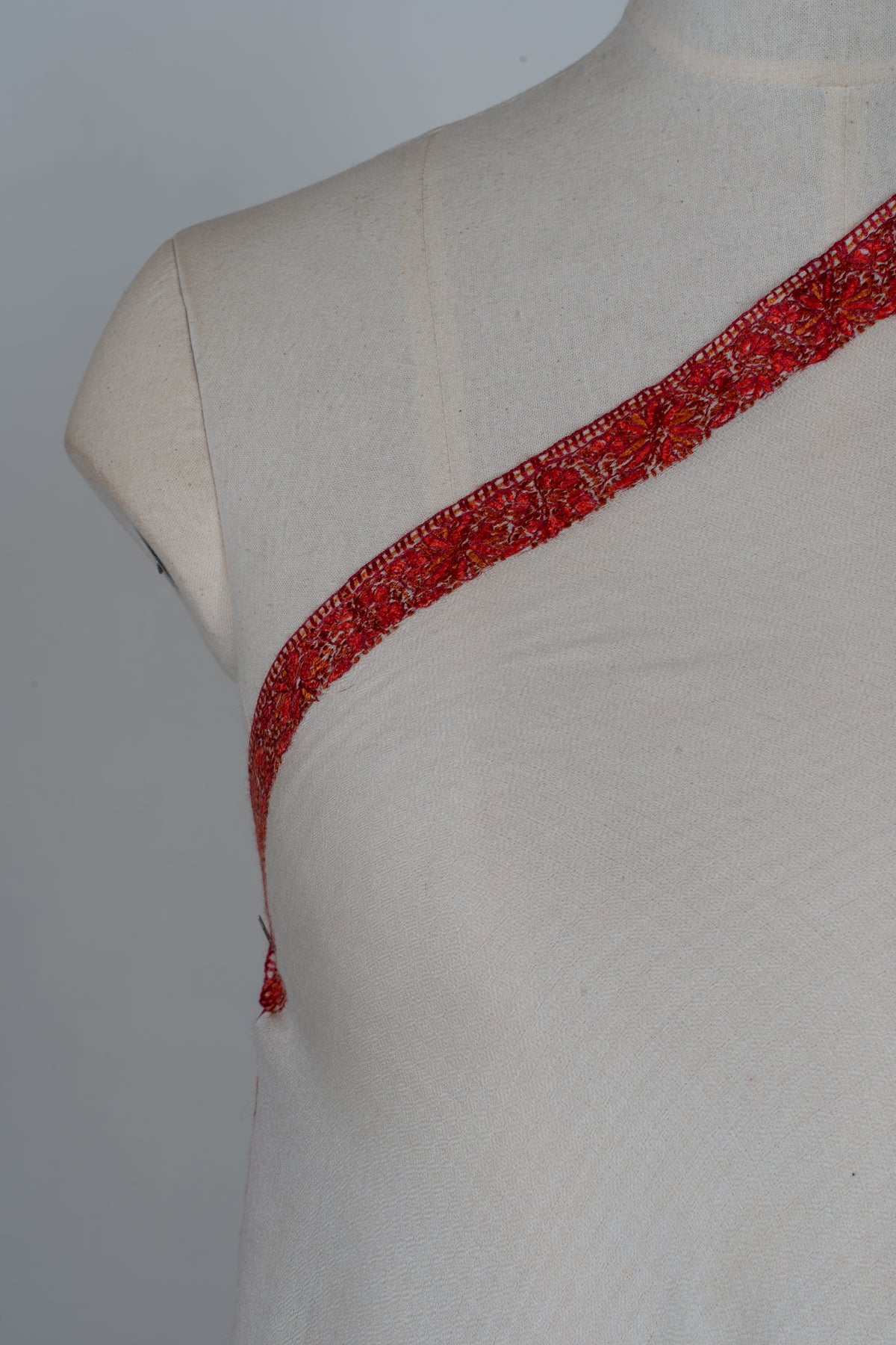 Chest detail of an ivory shawl with a narrow crimson floral border running diagonally across the shoulder