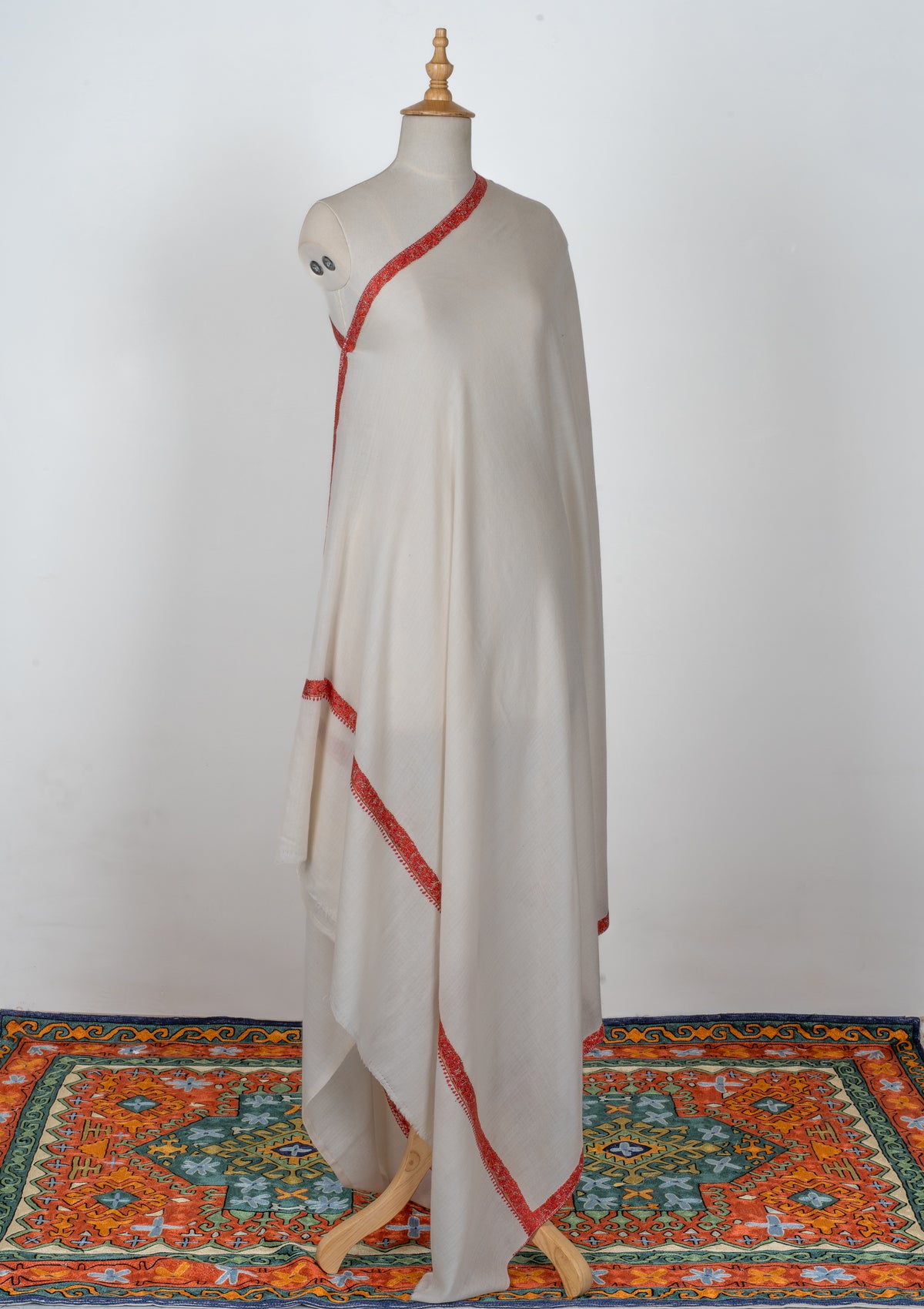 Full-length view of an ivory shawl draped over one shoulder of a mannequin, with narrow crimson embroidered borders