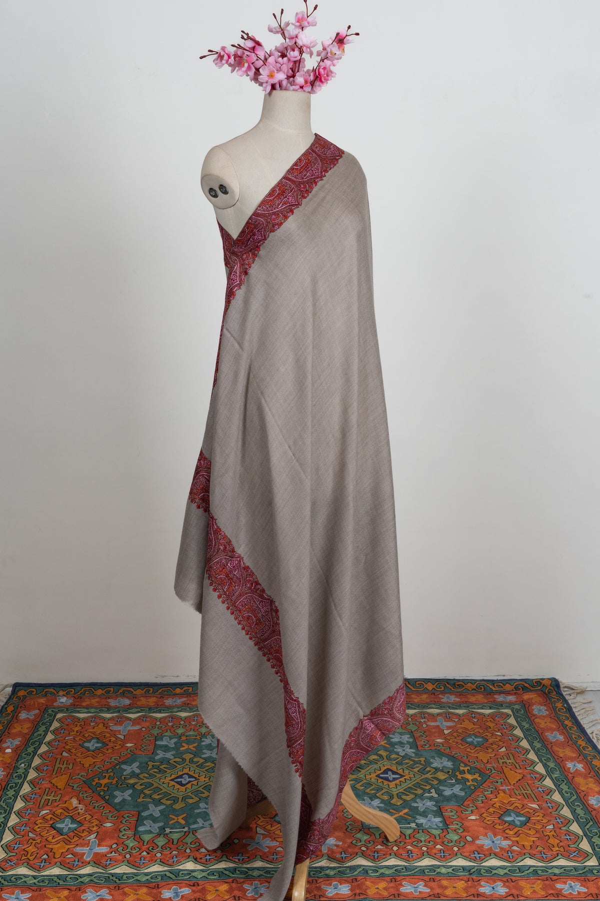 Full-length side view of an ash-grey merino wool shawl on a mannequin, edged with crimson patterned embroidery.