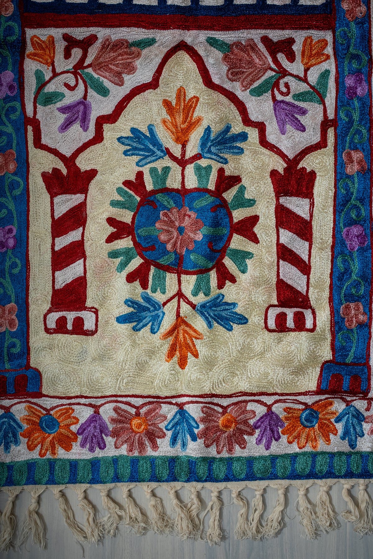 Lower end of the prayer mat with a blue floral circle between striped burgundy columns, flower borders and cream tassels