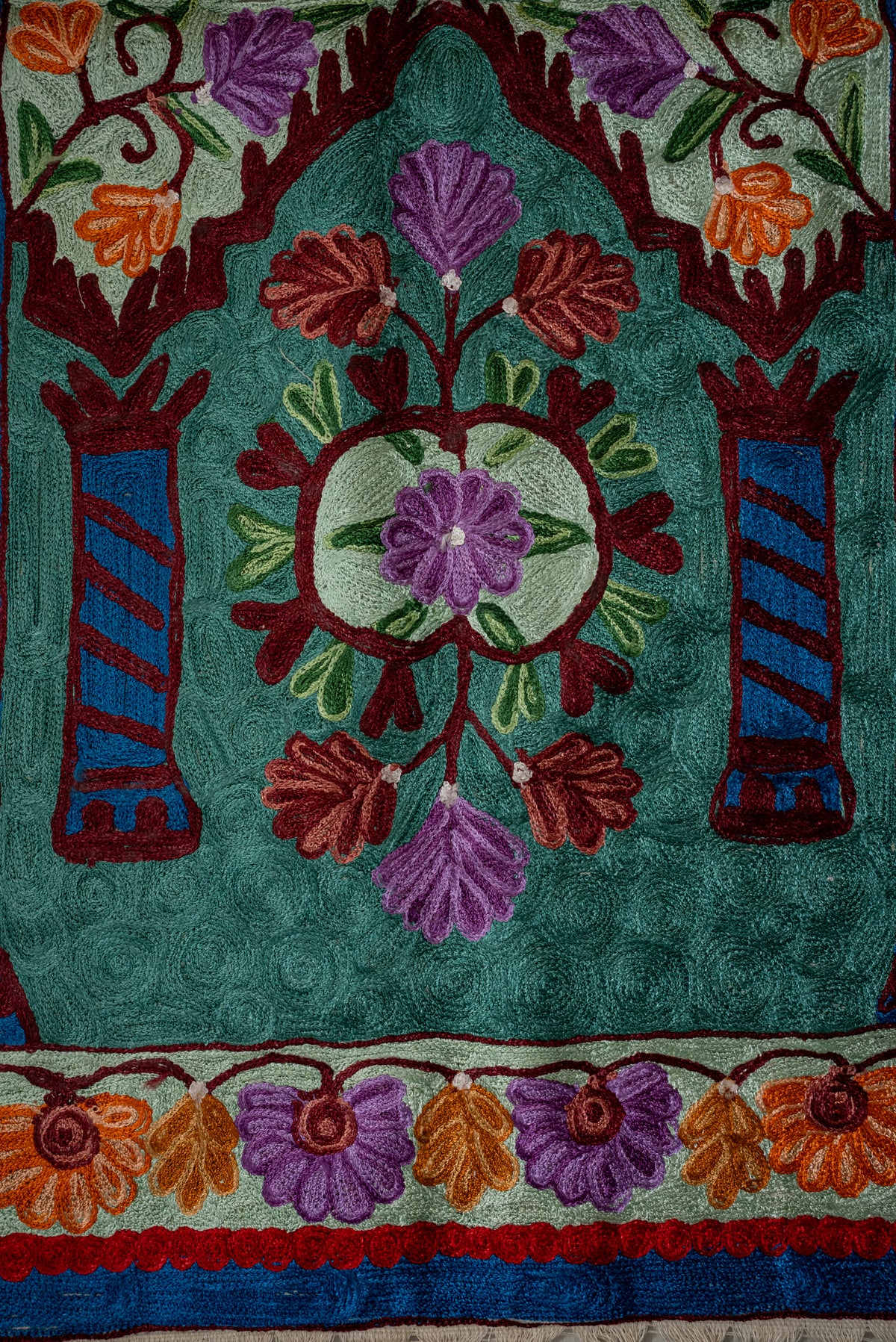 Embroidered floral medallion between blue columns under a burgundy arch on a teal prayer mat