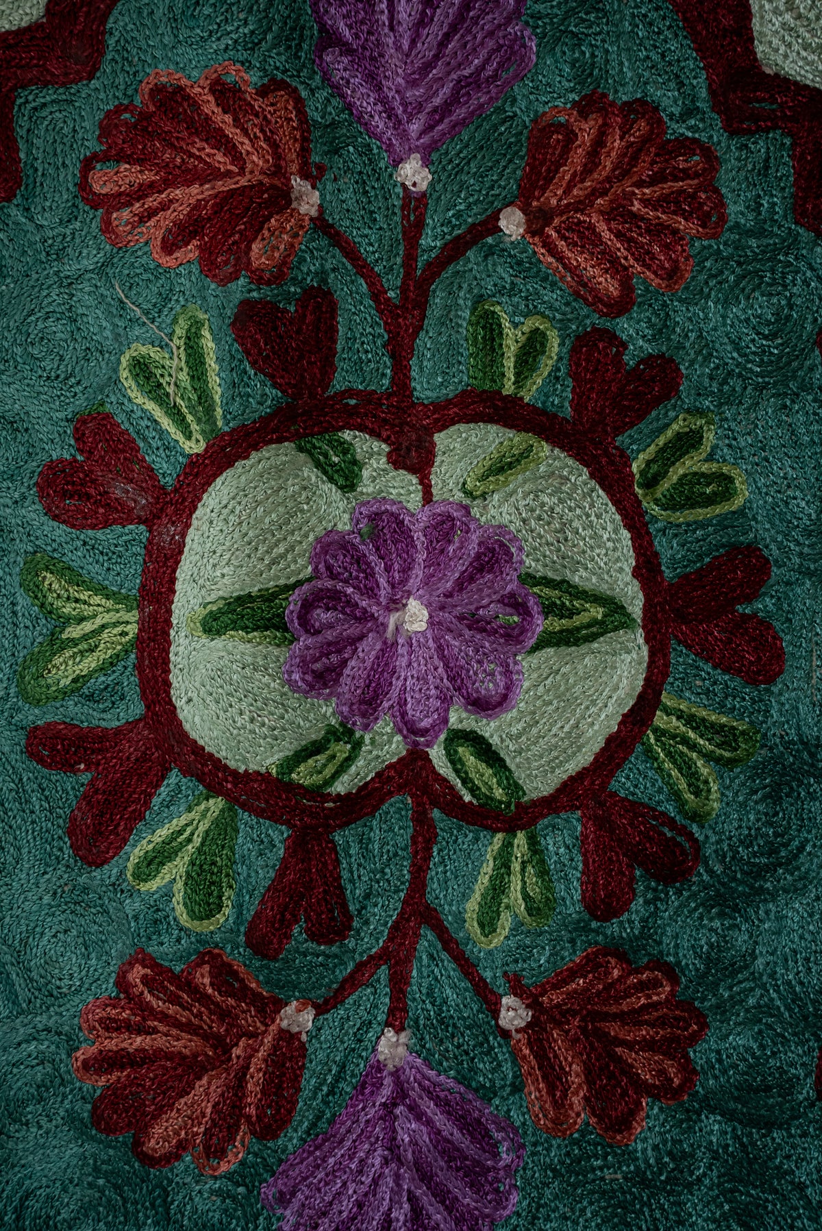 Close-up of a purple flower in a pale green medallion, with burgundy stems and rust-purple leaves on teal embroidery