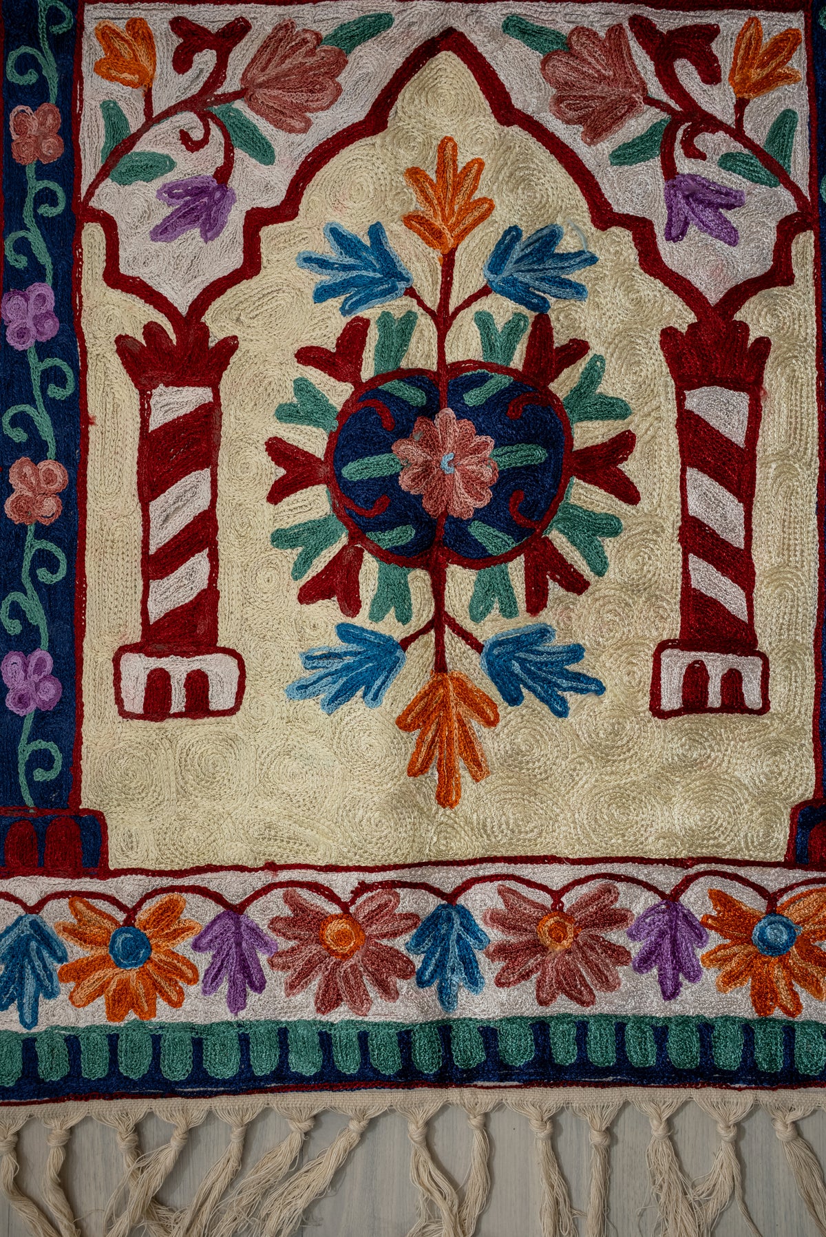 Central floral medallion between striped red columns, above a colourful flower border and cream tassels