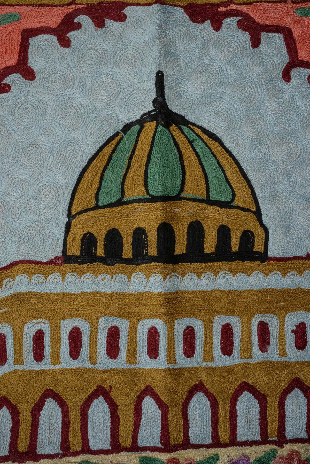 Close-up of a green-and-gold dome and rows of arches against the pale blue upper panel of an embroidered prayer mat