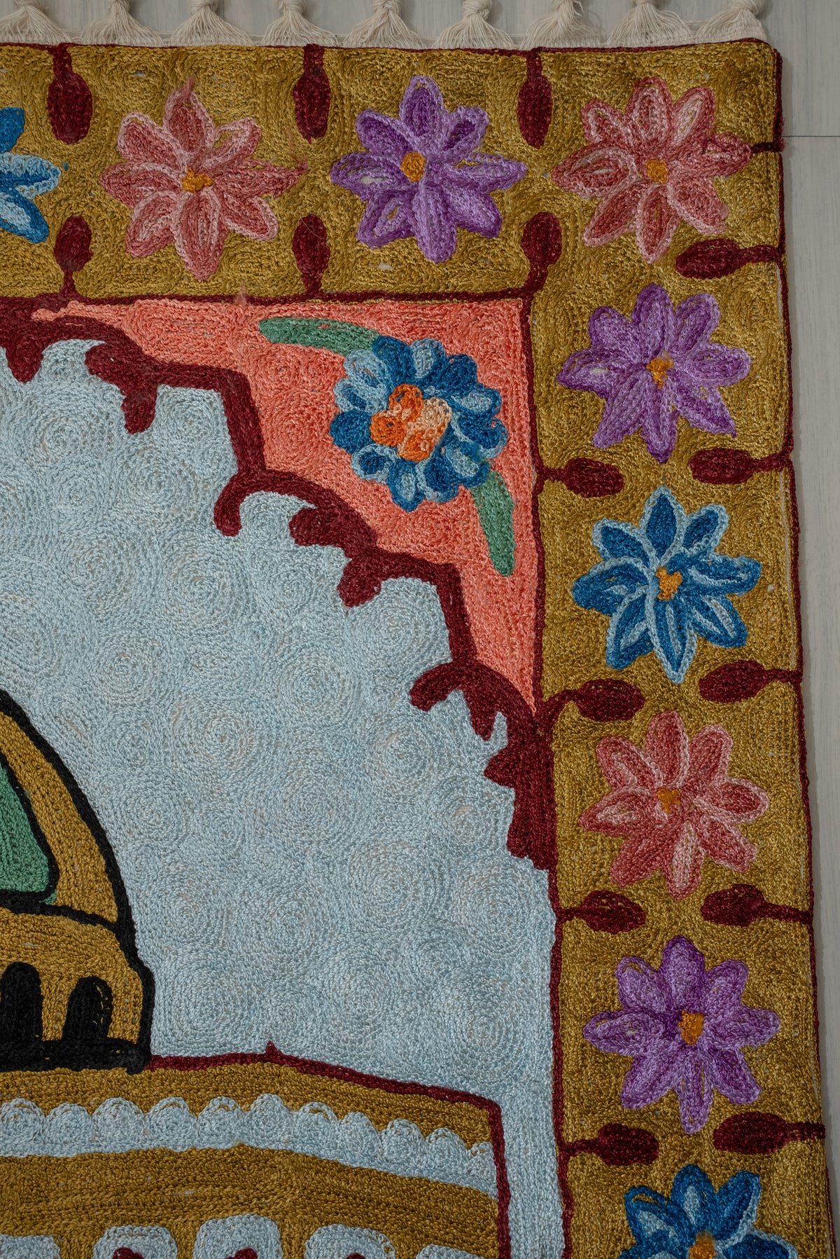 Upper corner of an embroidered prayer mat with pink, purple and blue flowers on ochre beside a pale blue arched panel