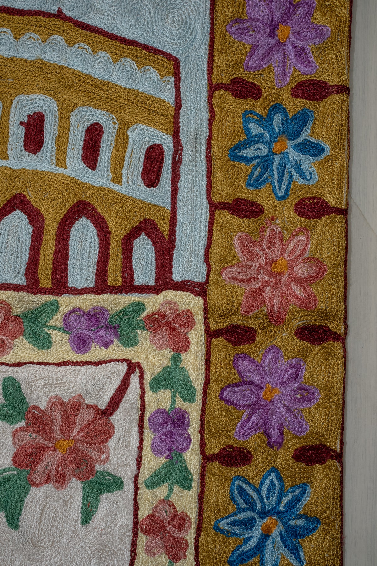 Close-up of pink, purple and blue flowers in an ochre prayer mat border beside the embroidered building and floral centre