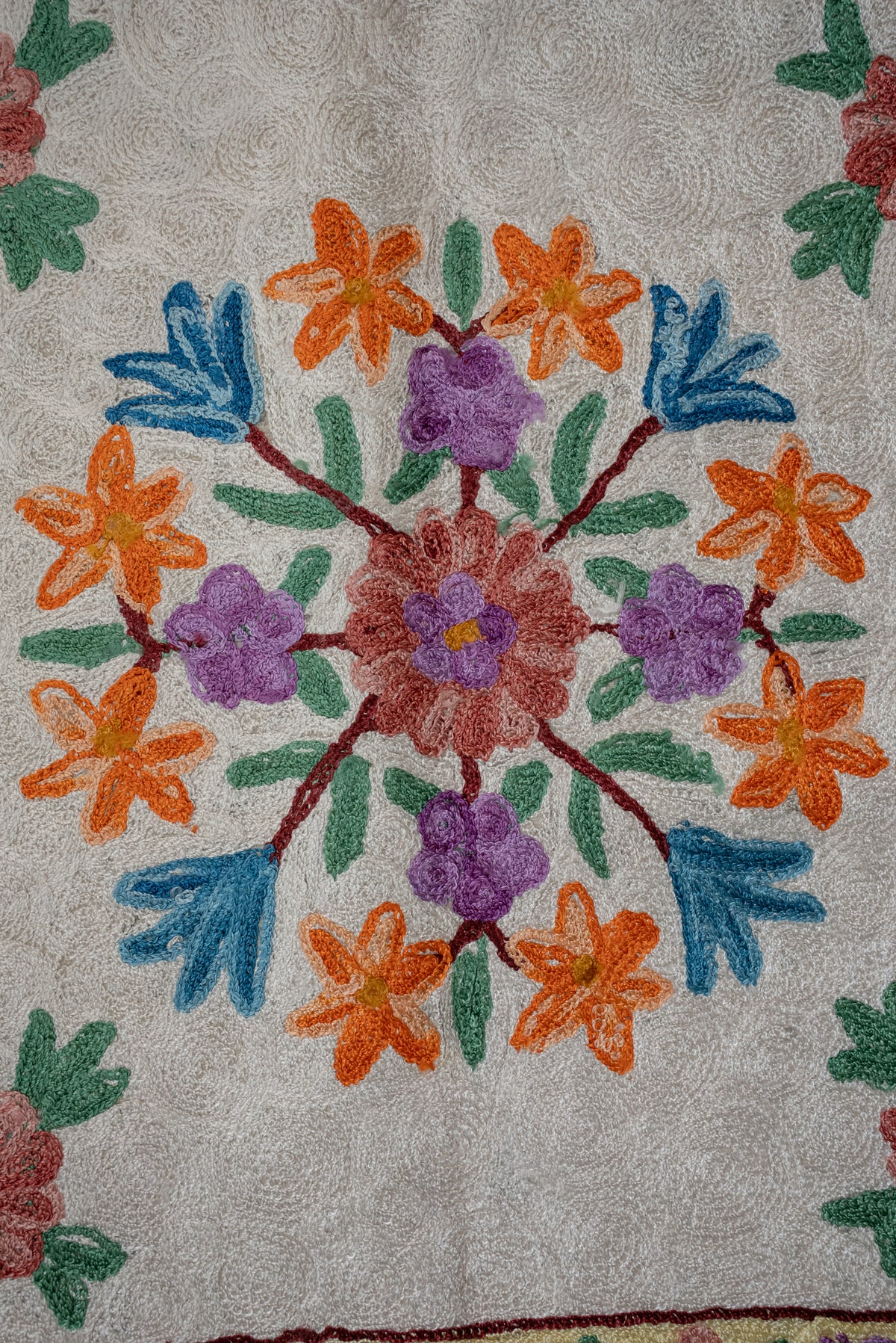 Close-up of orange, purple and pink flowers with blue and green leaves in the cream centre of an embroidered prayer mat