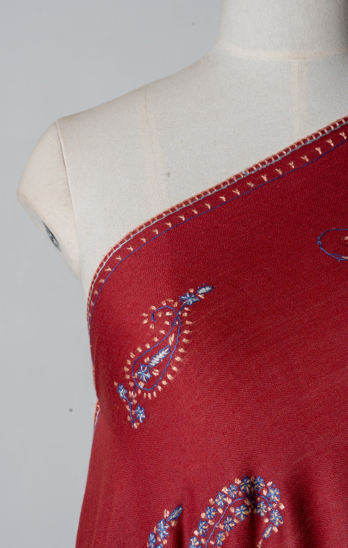 Shoulder detail of a deep red shawl with a small blue, pink and gold Sozni paisley beside narrow embroidered edging