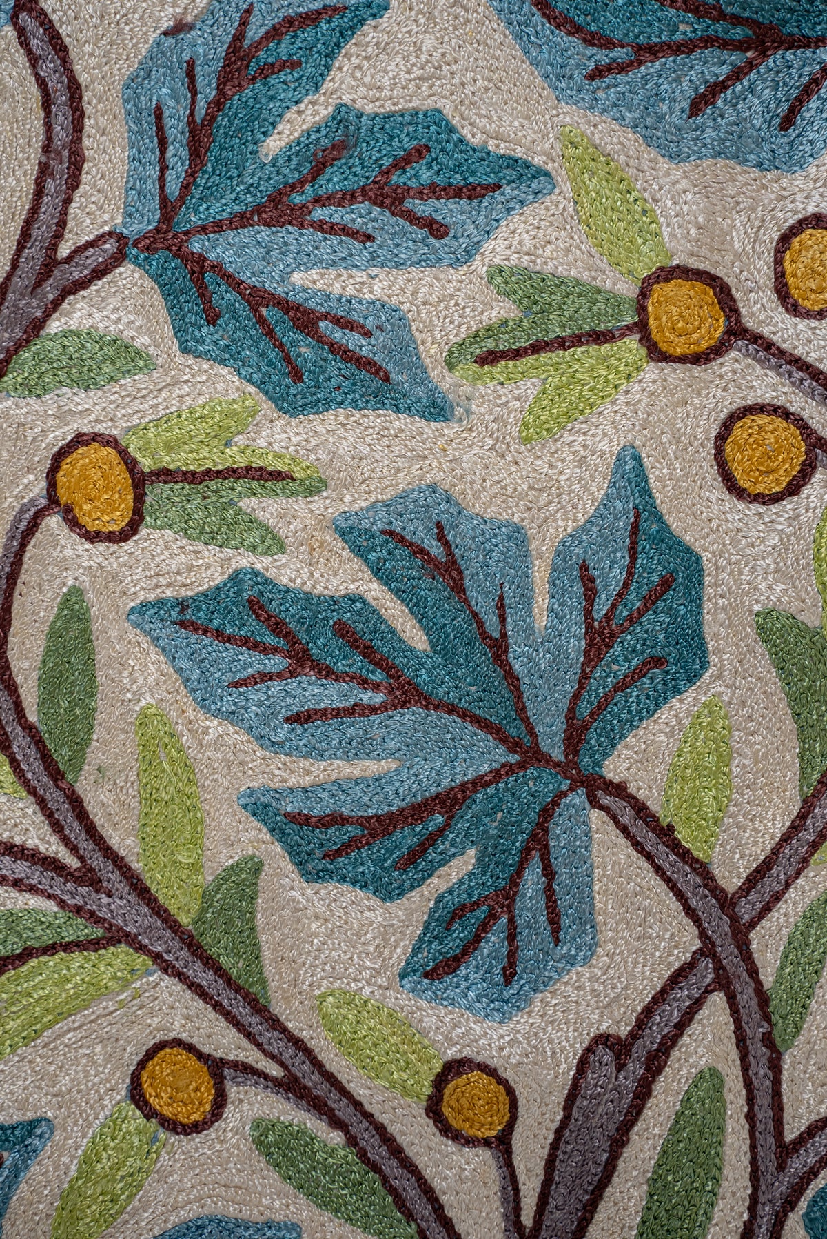 Close-up of teal leaves, green shoots and golden berries in the chain-stitch embroidery of an ivory rug