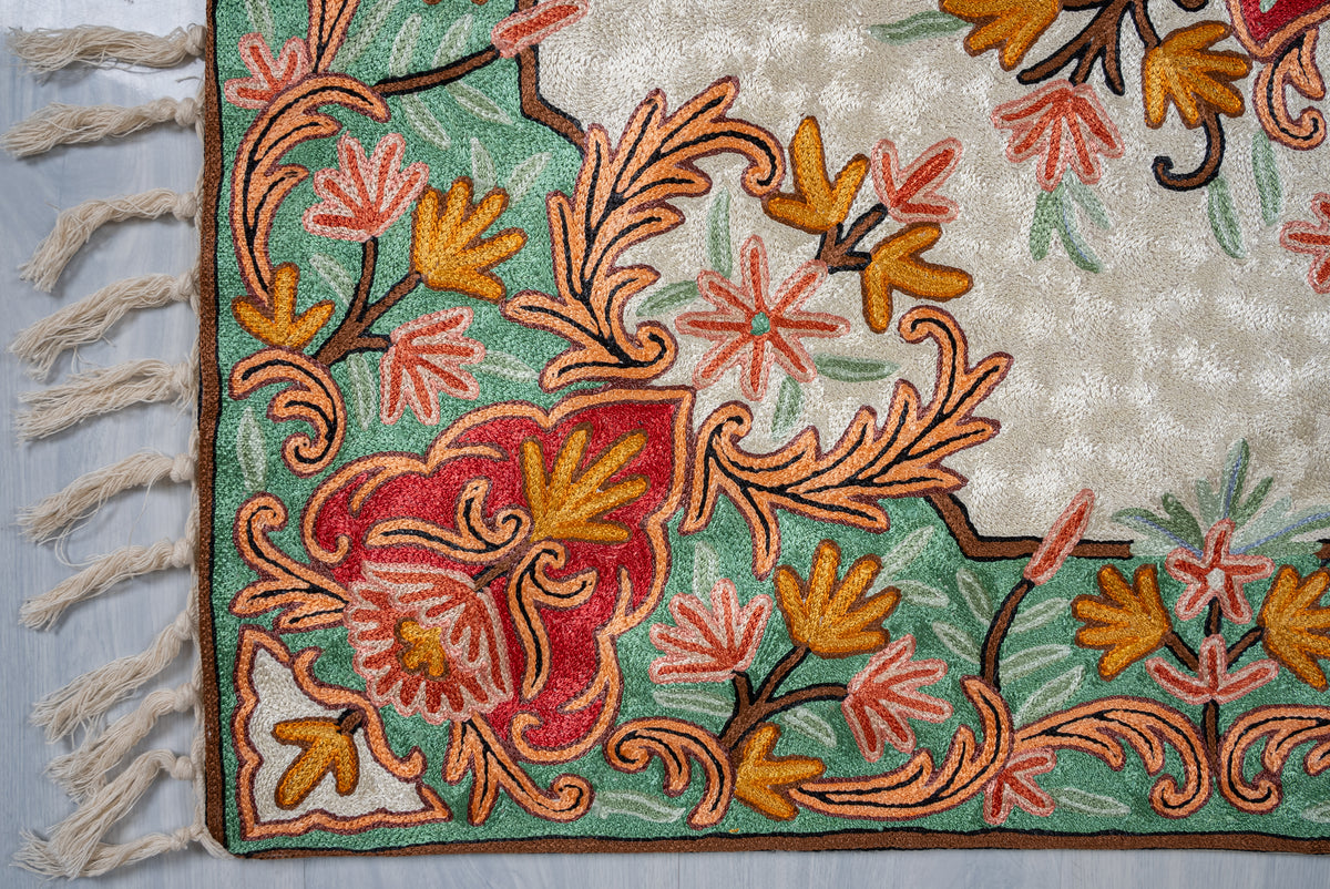 Corner of an Aari chain-stitch rug with coral and gold flowers, peach scrolling leaves, a green border and cream tassels