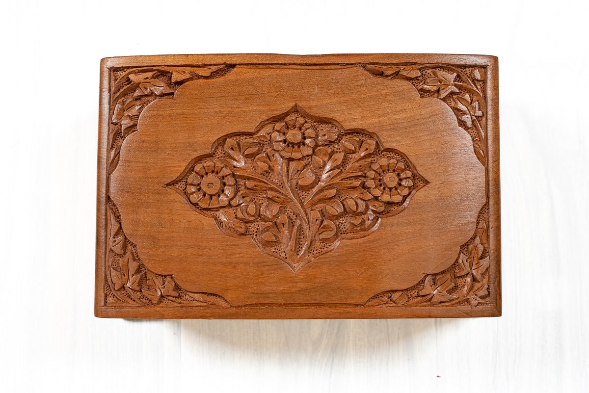 Top view of a walnut wood box lid with a carved central floral medallion and leafy corner motifs on smooth wood