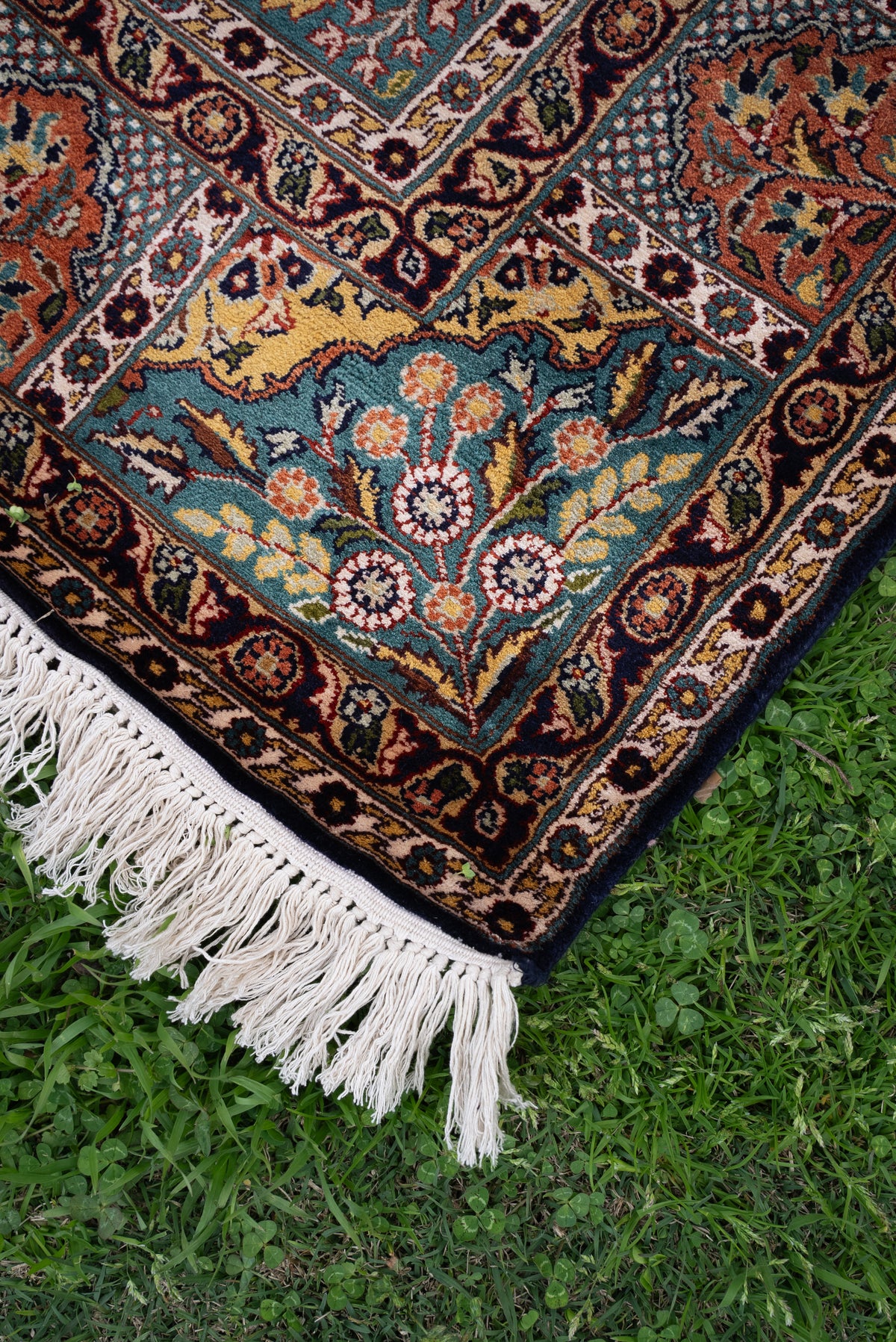 Angled corner of a silk carpet on grass, showing framed floral panels, dark patterned borders and cream fringe