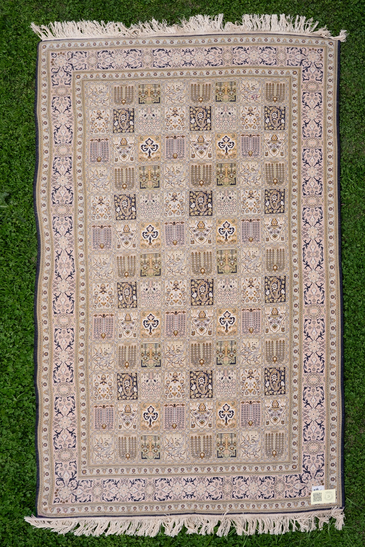 Ivory silk carpet laid flat on grass, with a grid of small patterned panels, navy floral borders and pale fringe