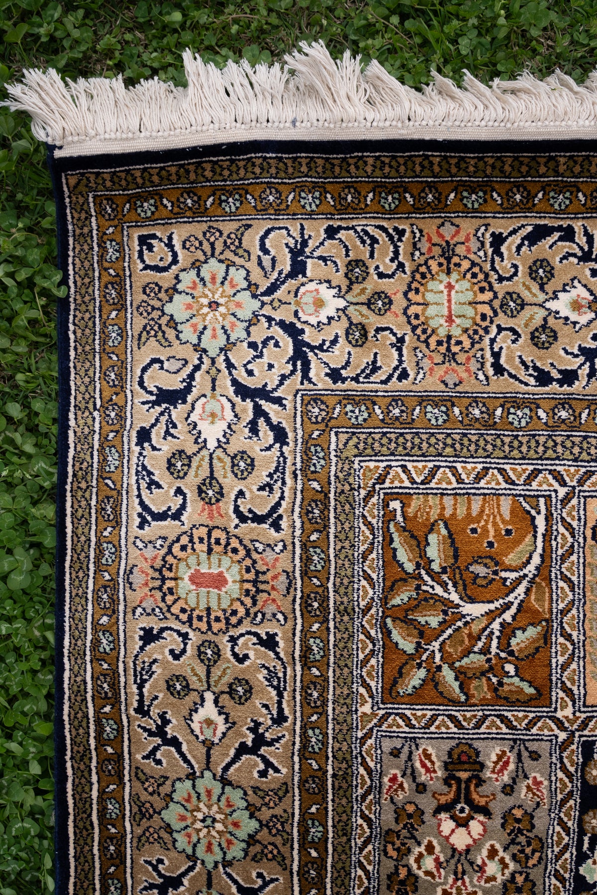 Corner detail of a silk carpet showing navy floral vines on ivory, gold patterned bands and white fringe