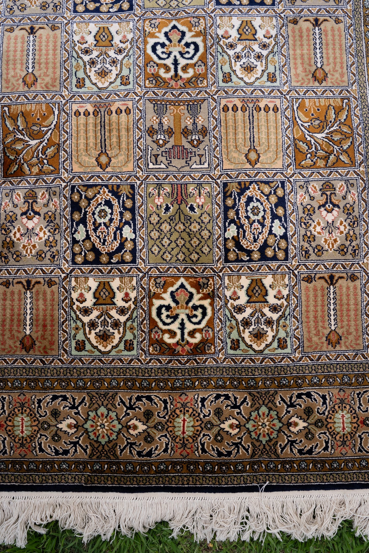 End section of a silk carpet with rows of ornamental panels above a broad ivory floral border and white fringe