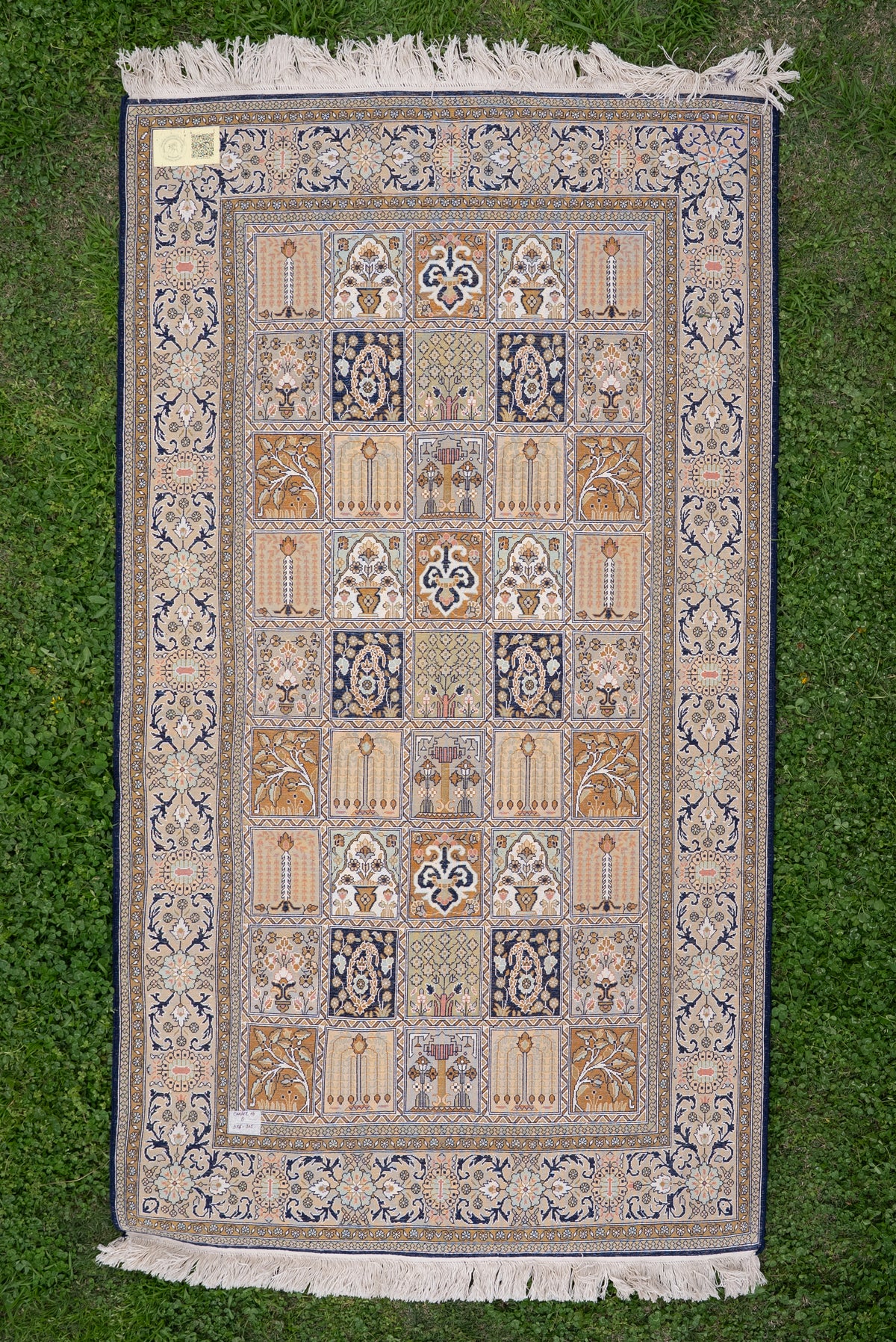 Ivory, navy and gold silk carpet laid flat on grass, with a grid of ornamental panels, floral borders and white fringe