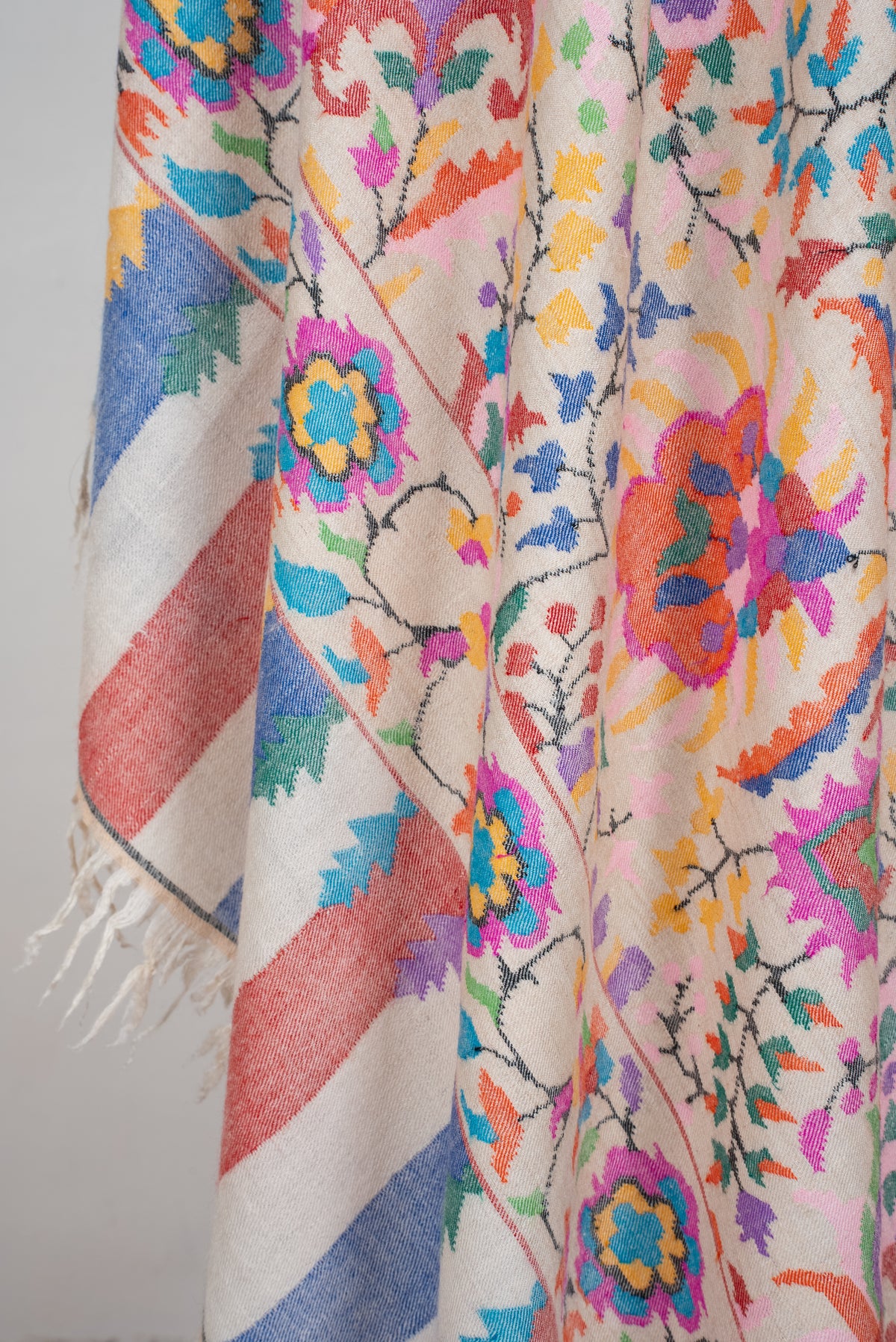 Close-up of ivory Pashmina shawl folds with colourful floral motifs, blue and pink end stripes and cream fringe