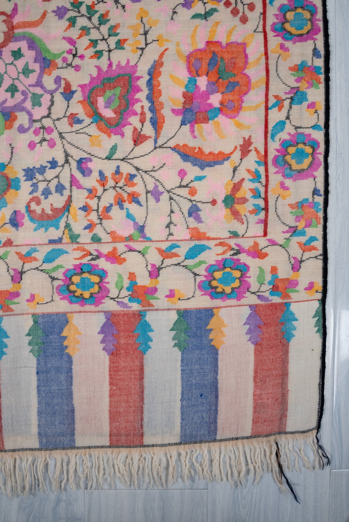 Corner detail of ivory Pashmina shawl with colourful flowers, a dark outer edge, striped end panels and cream fringe