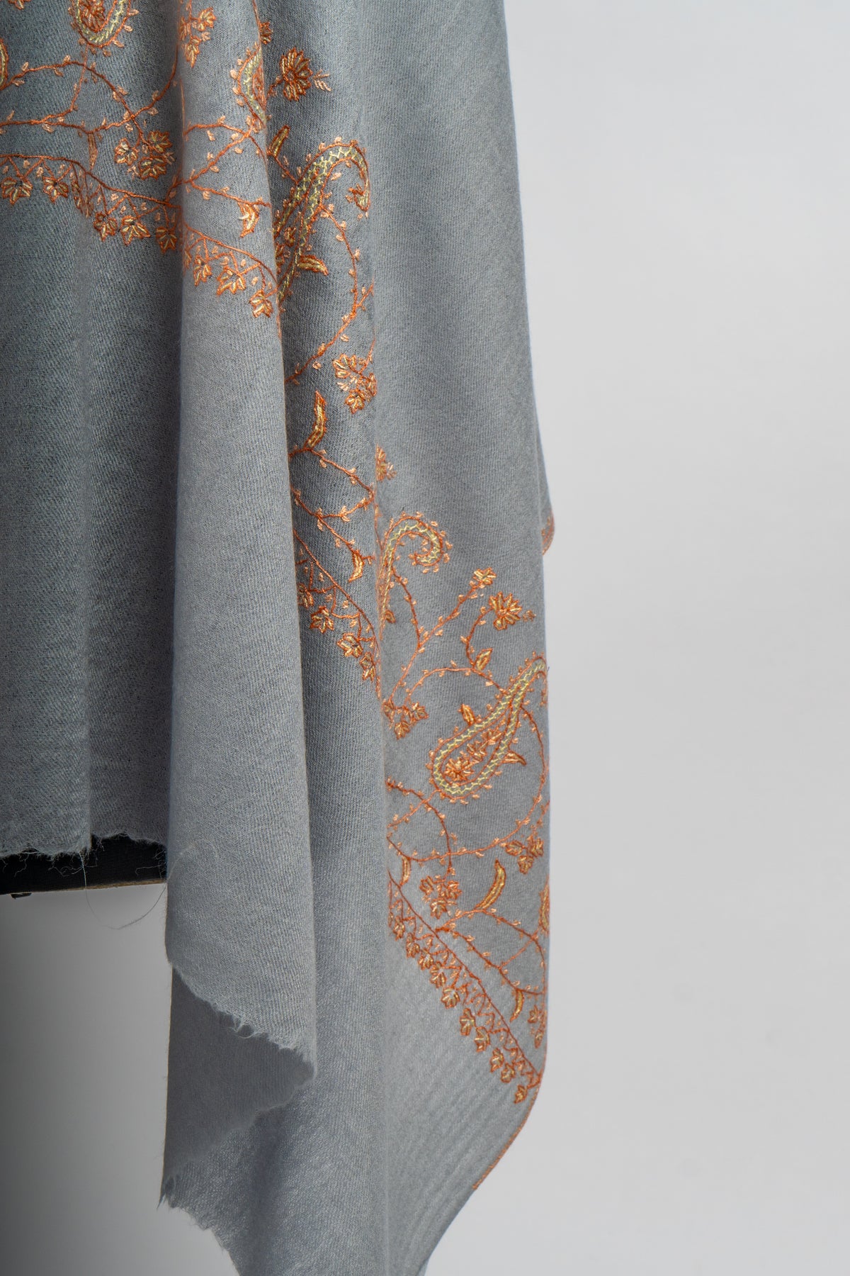 Hanging corner of a grey stole with delicate orange paisley and floral embroidery above a softly fringed edge.