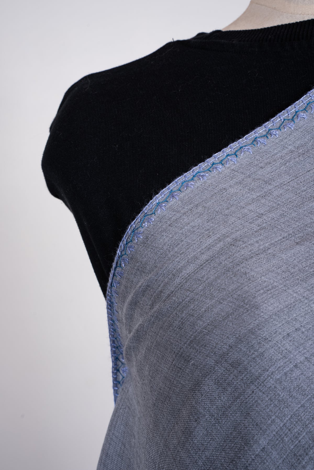 Close-up of a powder-blue merino wool scarf across a mannequin's chest, showing its fine texture and narrow blue embroidered edge.
