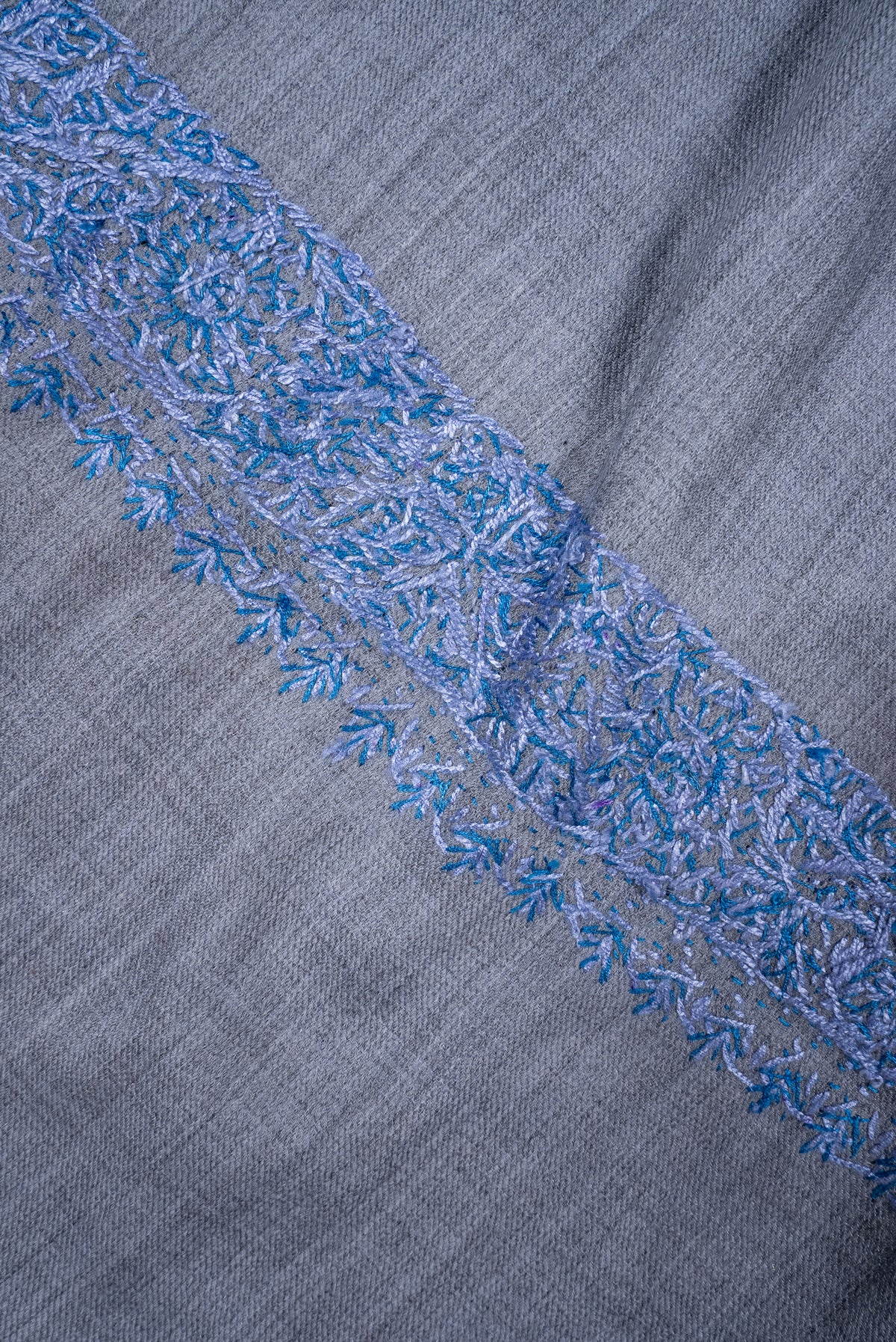 Close-up of blue embroidered floral vines and small leaf shapes in a border on a powder-blue merino wool scarf.