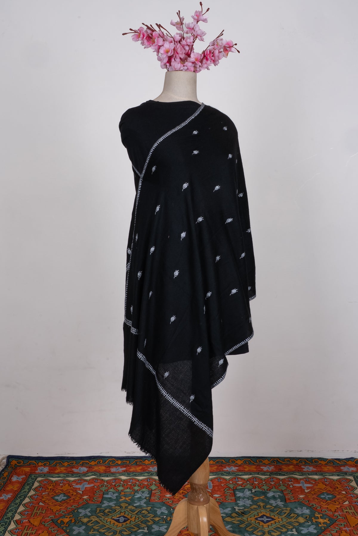 Black scarf draped on a mannequin, with scattered white embroidered floral motifs and a fine leafy border above a plain black end.