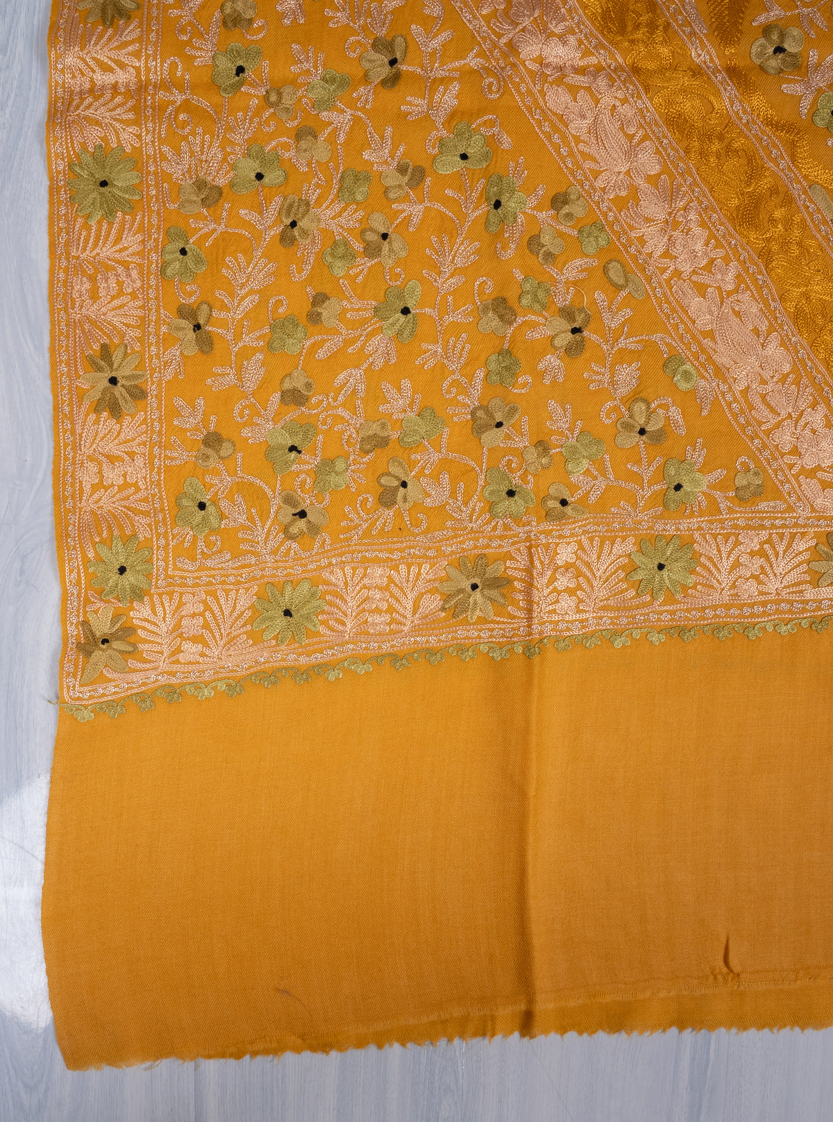 Flat corner detail of the mustard shawl, with small olive-green Aari flowers and pale leafy borders above the plain end