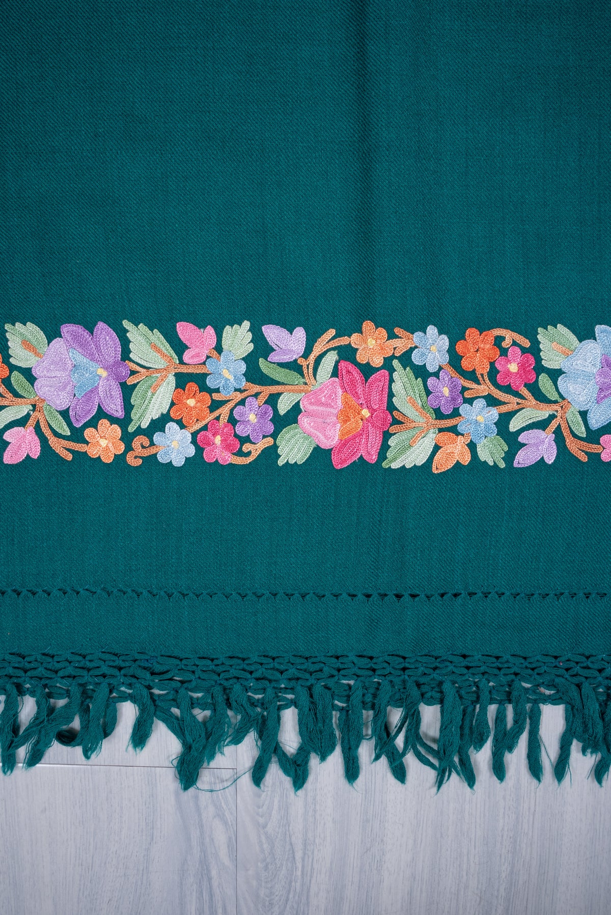 Flat detail of a teal shawl end, with a pastel Aari flower-and-leaf border above matching knotted fringe.