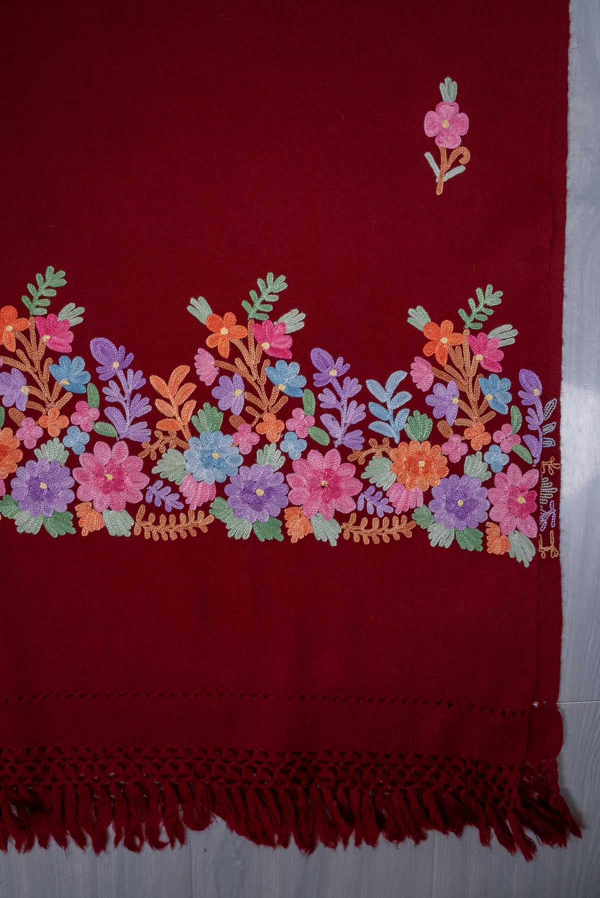 Corner of a maroon shawl laid flat, with a pink sprig above a multicoloured Aari floral border and red knotted fringe.