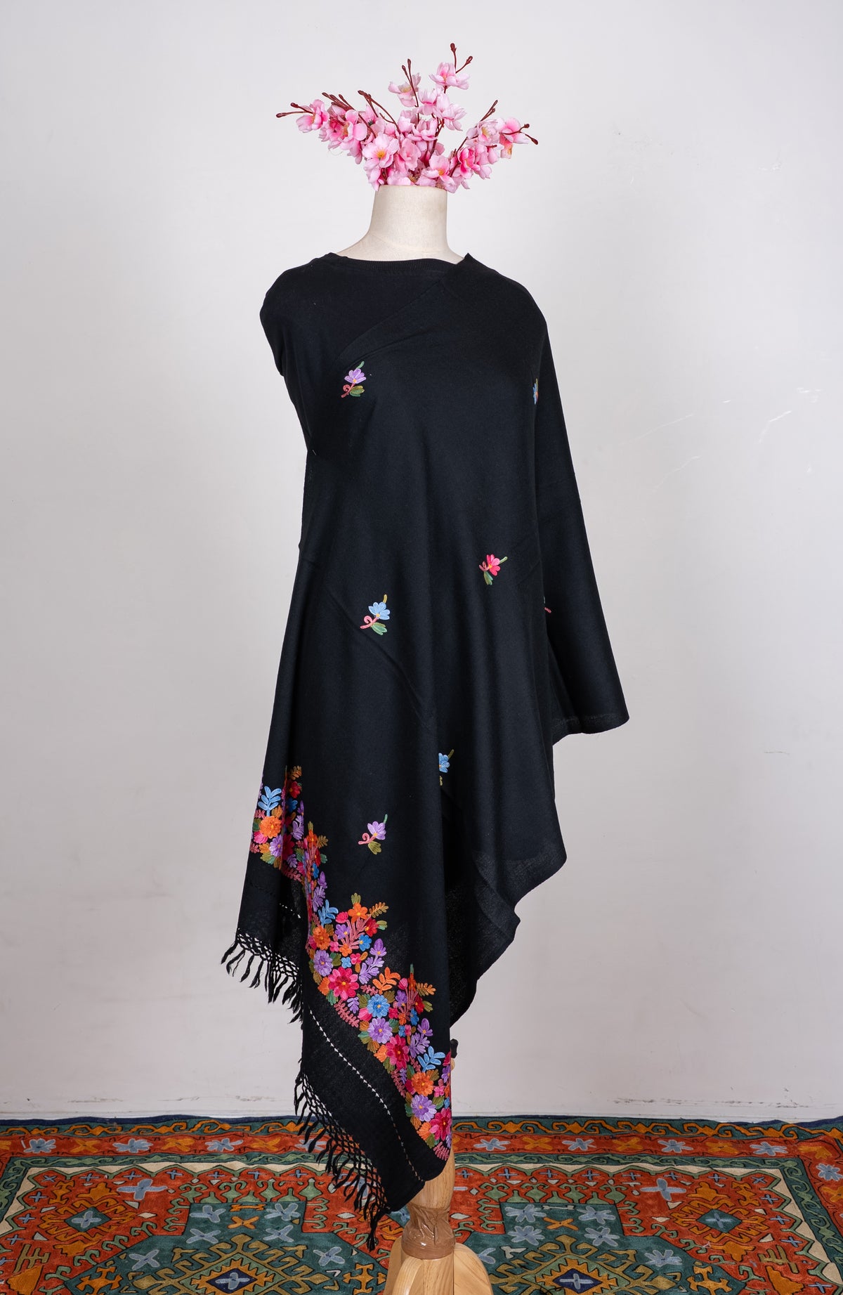 Black shawl draped around a mannequin, with scattered Aari floral sprigs and a bright multicoloured floral border above black fringe.