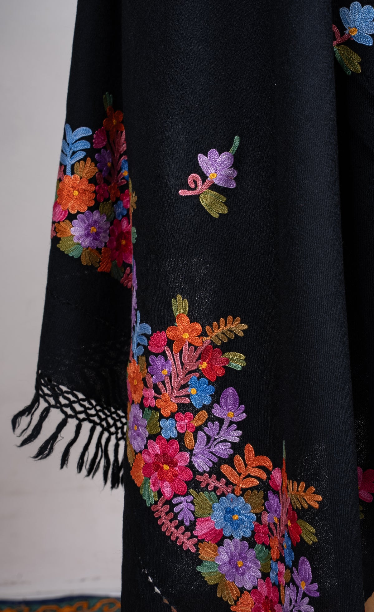 Close-up of a draped black shawl end, with bright orange, pink, blue and purple Aari flowers and knotted black fringe.