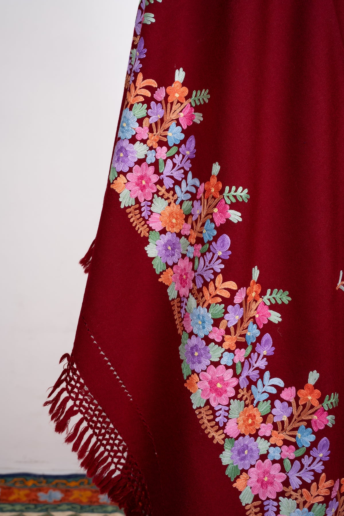 Close-up of a draped maroon shawl end, with pink, orange, blue and purple Aari flowers above knotted red fringe.