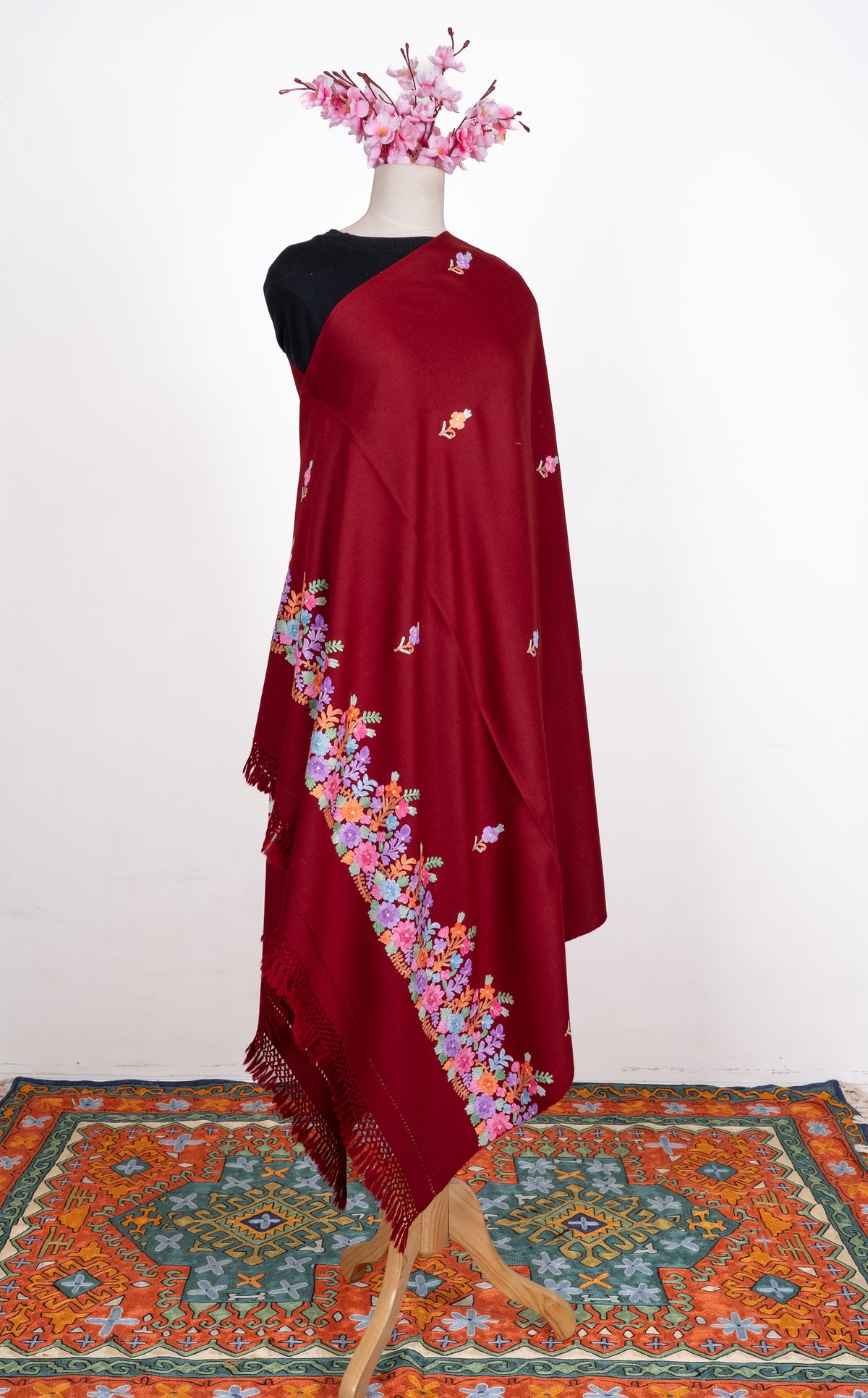 Maroon shawl draped on a mannequin, with scattered Aari floral sprigs, a bright multicoloured flower border and knotted fringe.