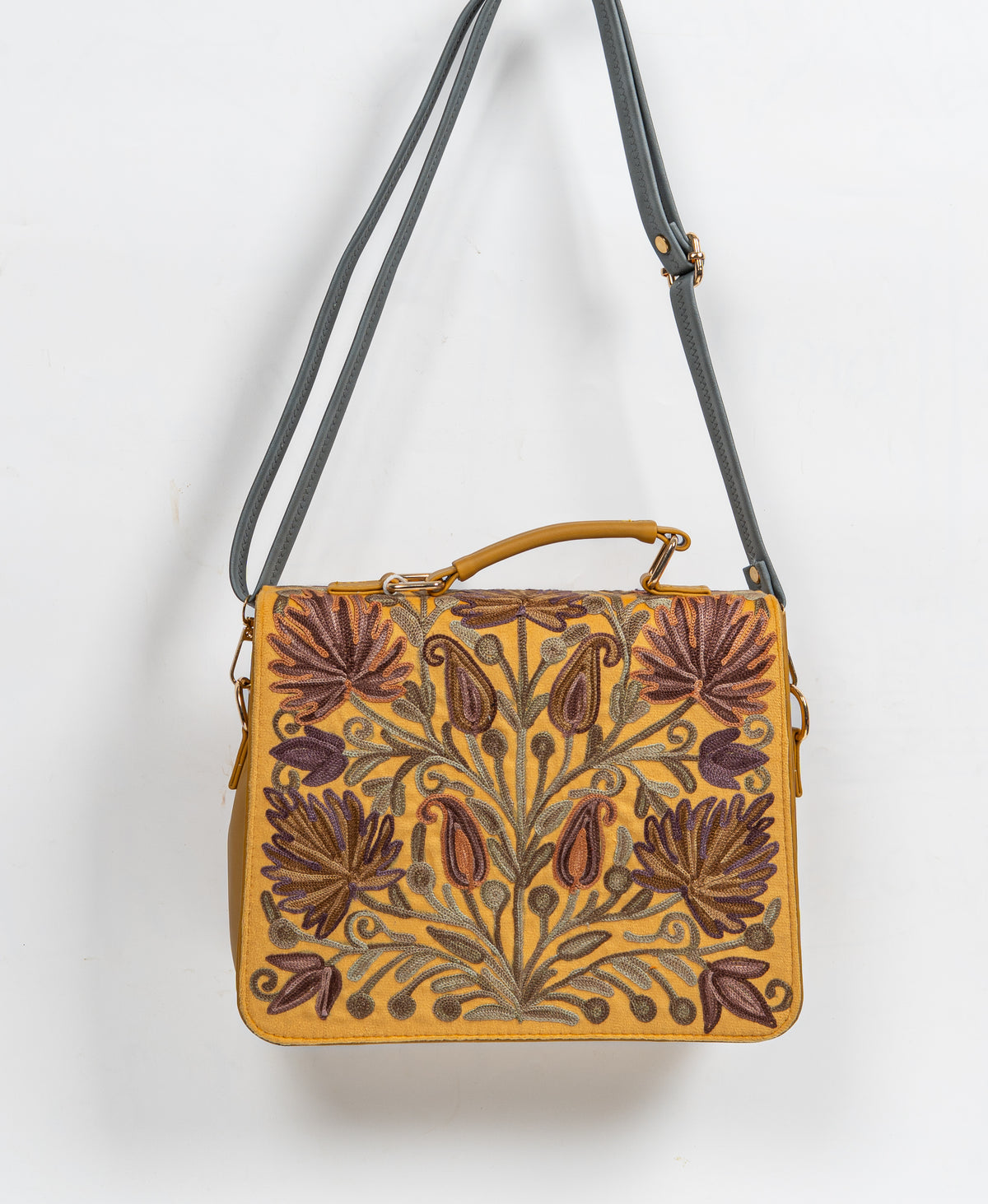 Mustard embroidered handbag hanging from a long grey shoulder strap, with its floral front flap facing forward.