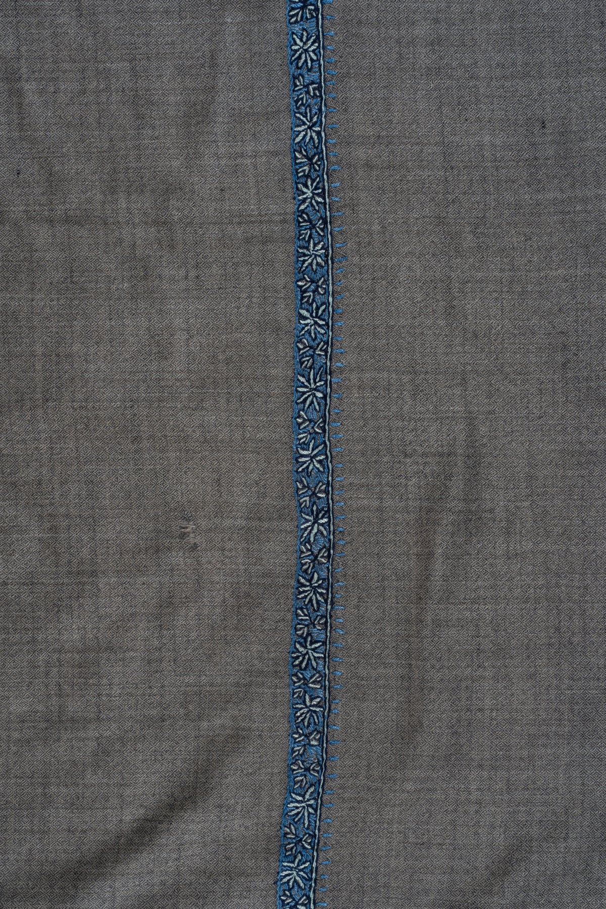 Close-up of a narrow blue Sozni border with pale flowers and leaves on grey Merino wool.