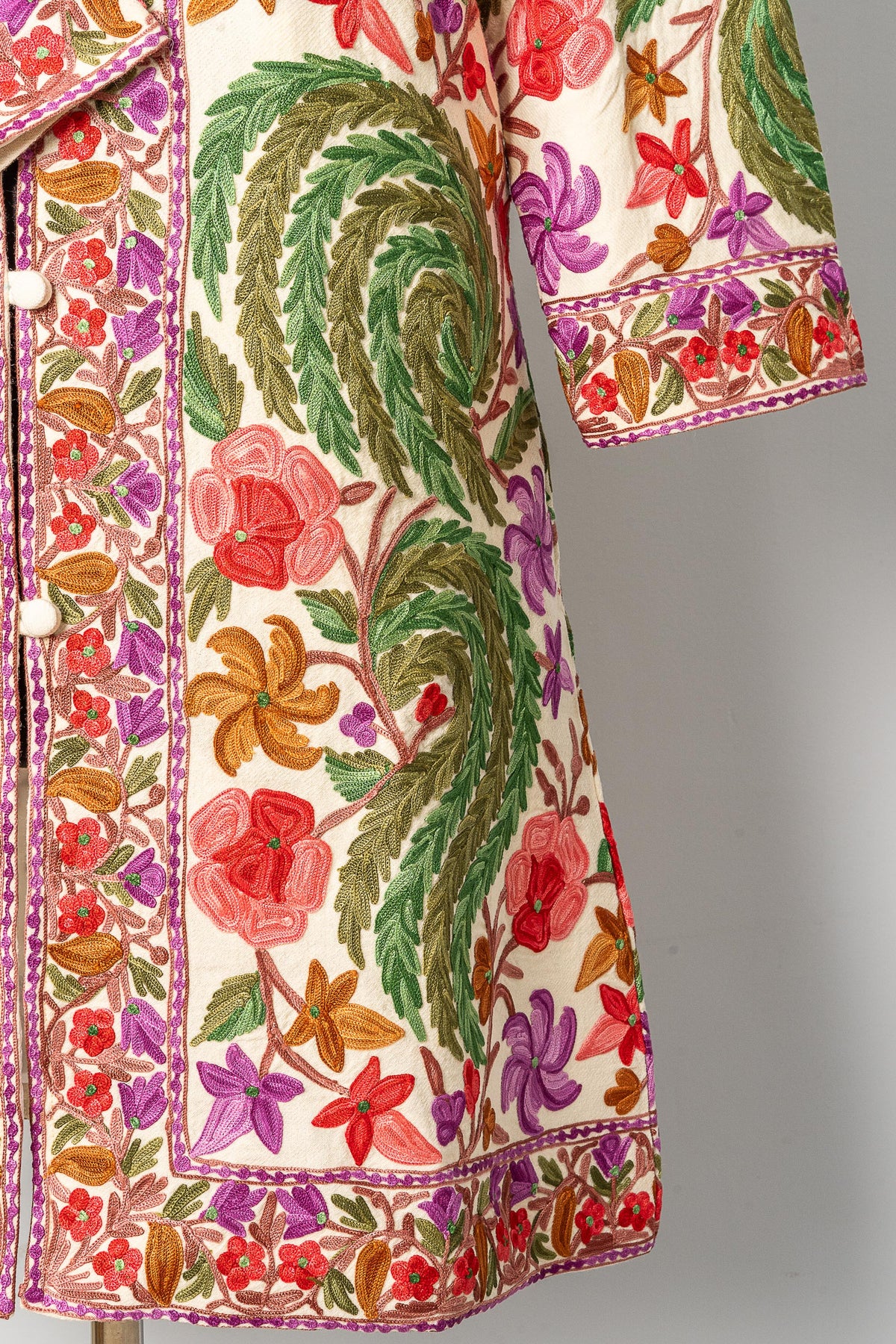 Lower-front detail of the white jacket, with green leafy embroidery, colourful flowers and a narrow floral hem border
