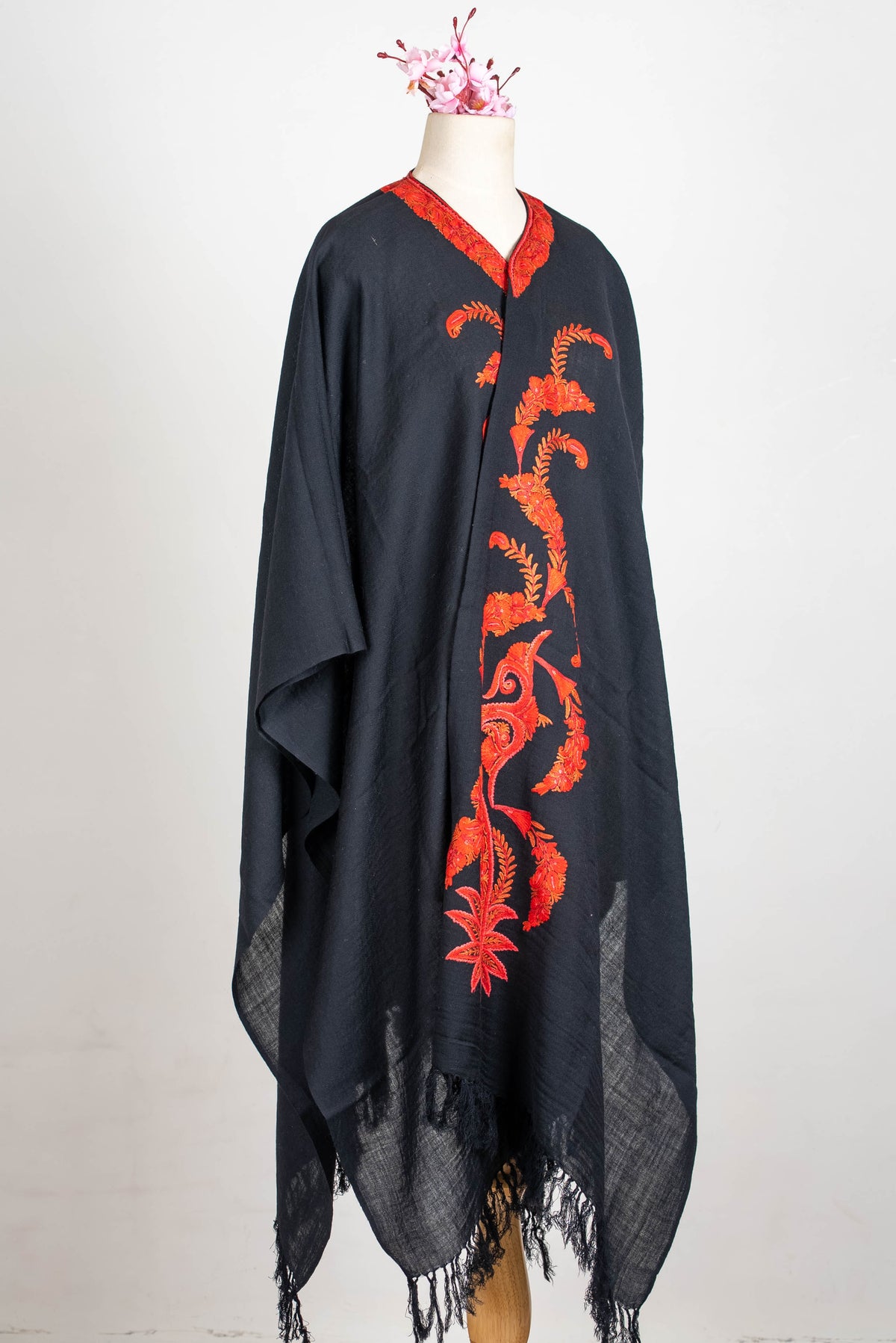 Full-length angled view of a black cape on a mannequin, with red-orange front embroidery and a fringed hem