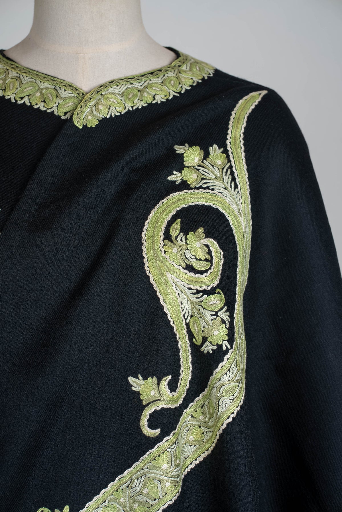 Close-up of sage-green and ivory paisley embroidery beside the black cape’s decorated neckline
