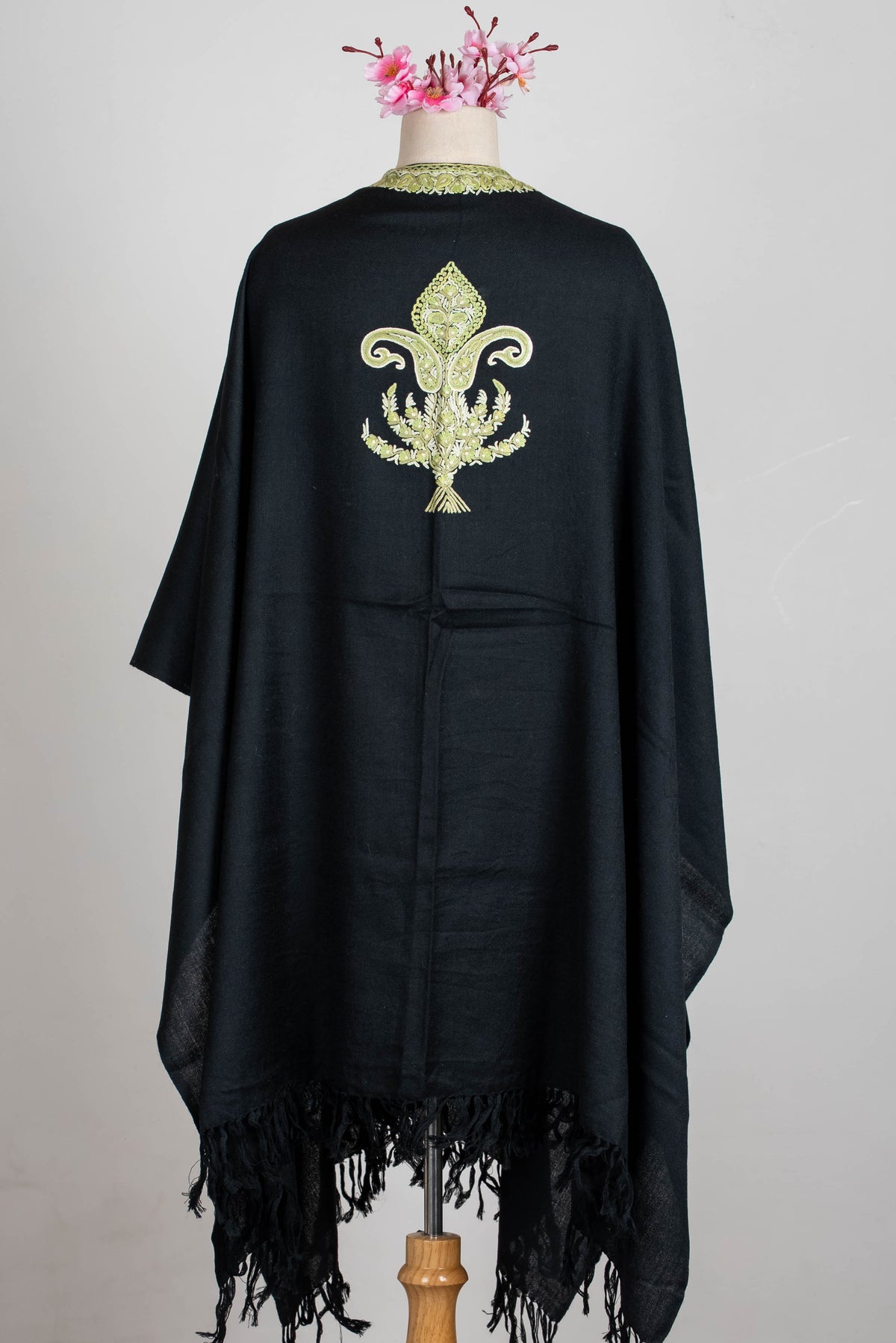 Full back view of the black cape on a mannequin, with one sage-green ornamental motif, embroidered neckline and black fringe