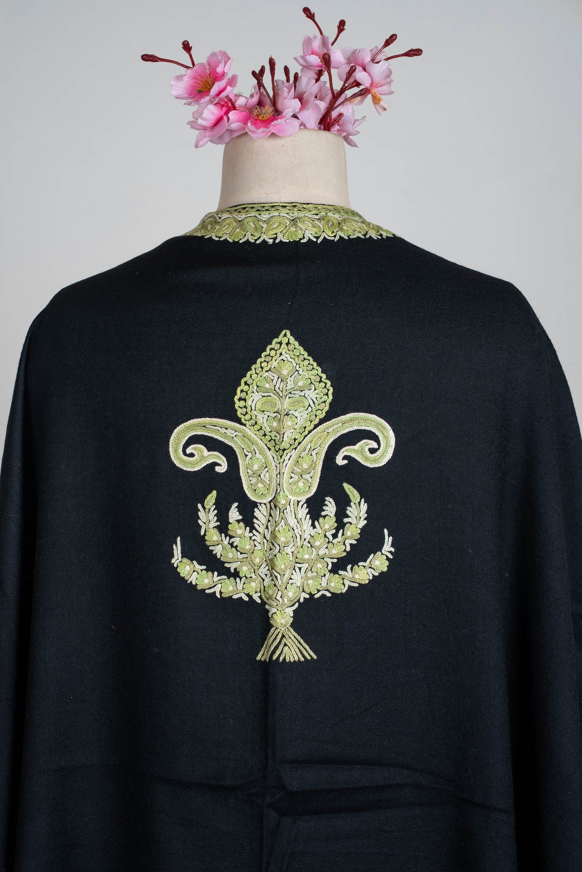 Upper-back detail of the black cape, showing a sage-green and ivory ornamental motif below the embroidered neckline