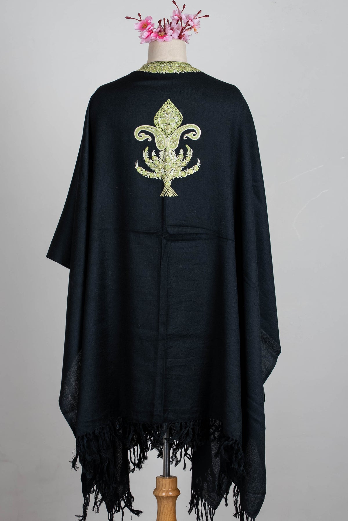 Straight back view of the black cape on a mannequin, with a single sage-green embroidered motif and long black fringed ends