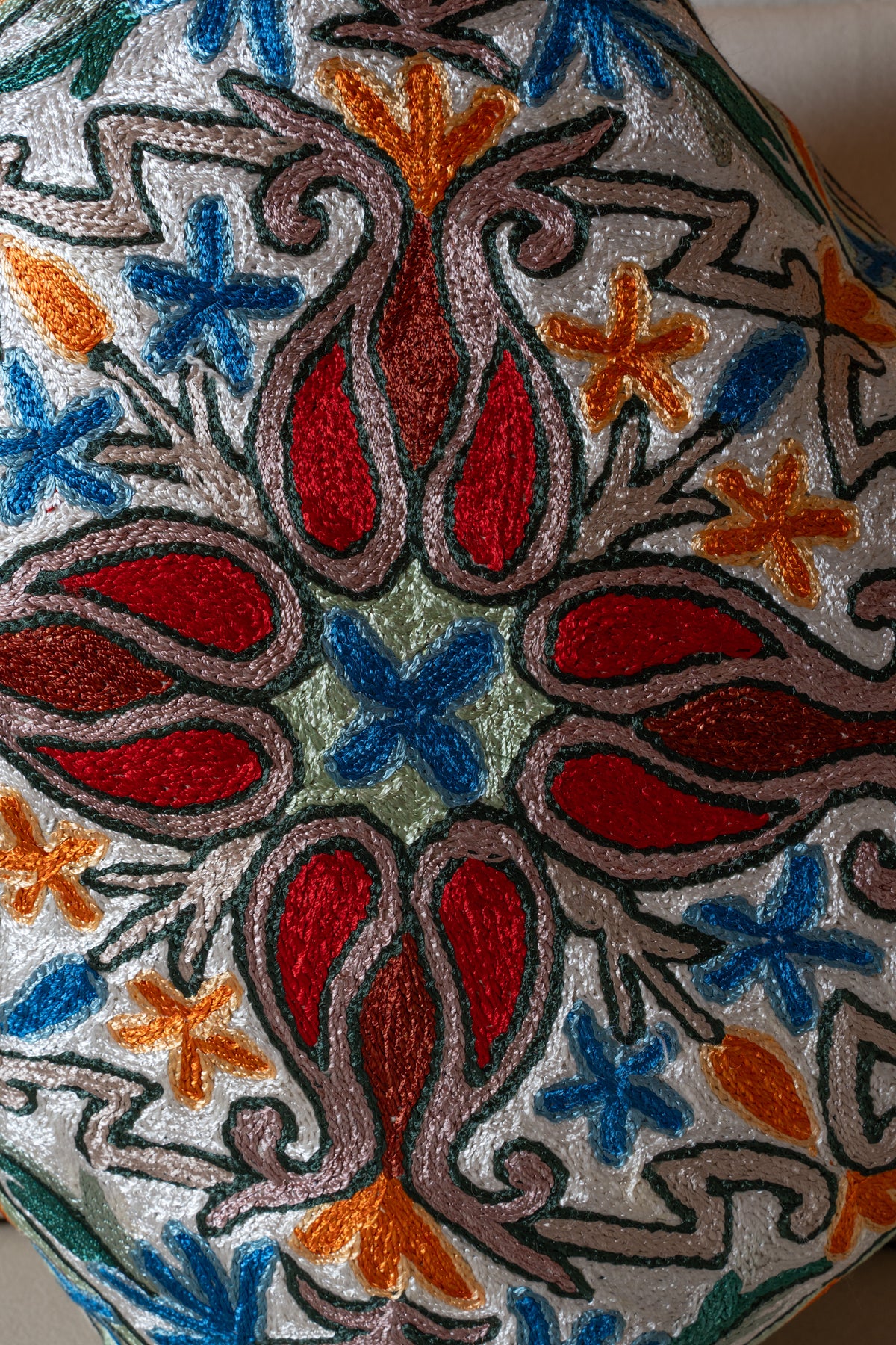 Close-up of red teardrop shapes, blue flowers and orange details in textured embroidery on an ivory cushion