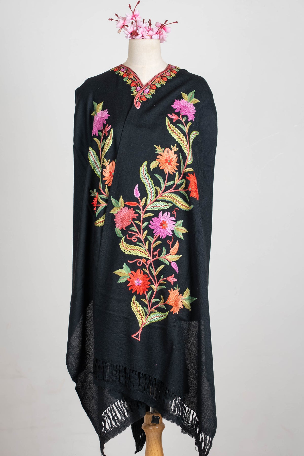 Long front view of the black shawl cape on a mannequin, with colourful botanical embroidery, a decorated neckline and black fringed ends